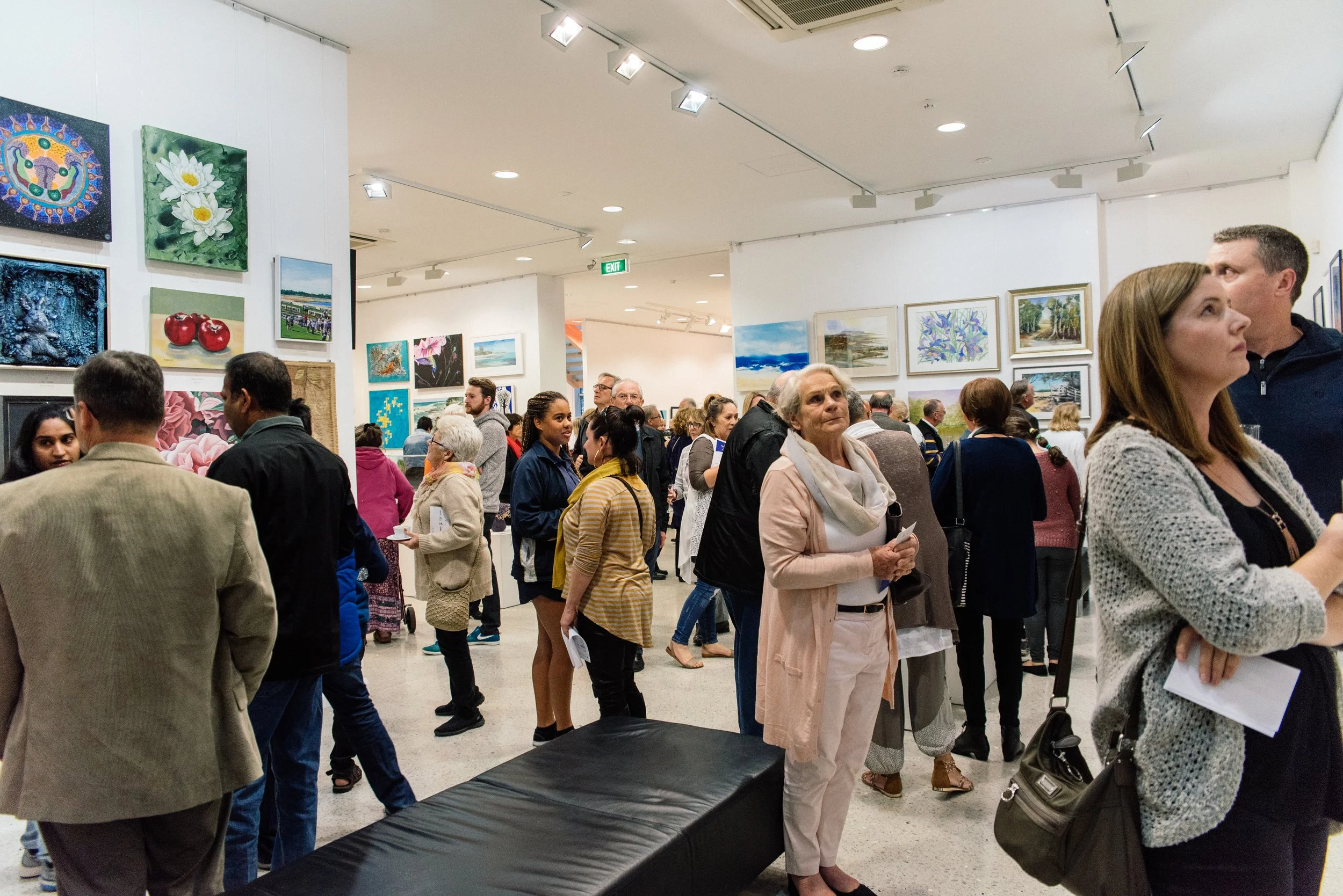 A gallery full of standing patrons. The walls are covered in art, and a woman stands in the foreground.