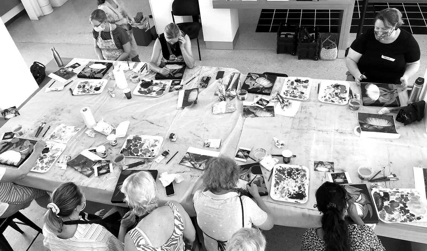 A black and white photograph of a top-down view. A table stretches across the room with several people creating art on it.