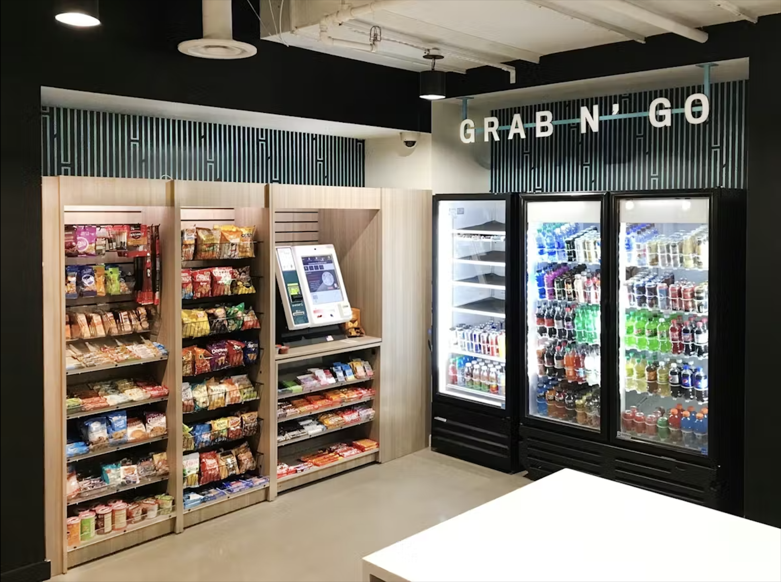 Snack and beverage vending area with snacks on wooden shelves, refrigerated drinks in glass-door coolers, and a self-service payment kiosk, under a sign that reads 'GRAB N GO' in a modern retail setting.
