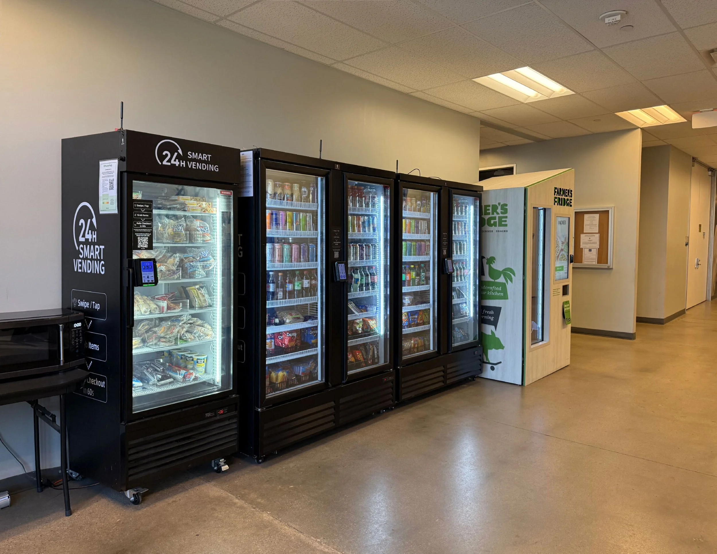 Lineup of four vending machines, the first black with a digital keypad, the next three glass-fronted with various snacks and drinks, located against a plain wall in a corridor.