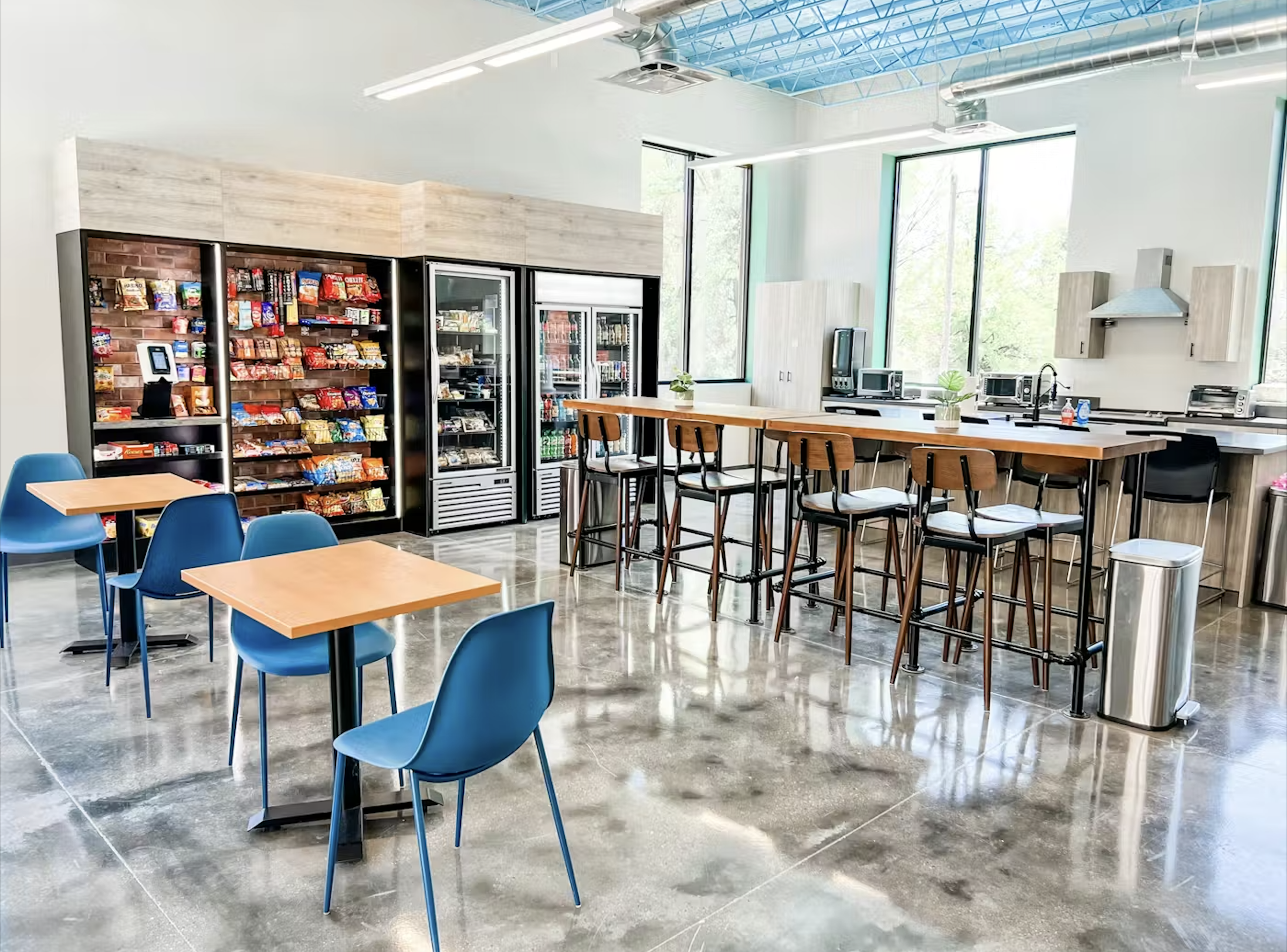 Modern break room with small square tables and blue chairs, a snack vending machine, refrigerated drinks, a high countertop with bar stools, microwaves, and large windows letting in natural light. Suitable for a casual office setting.
