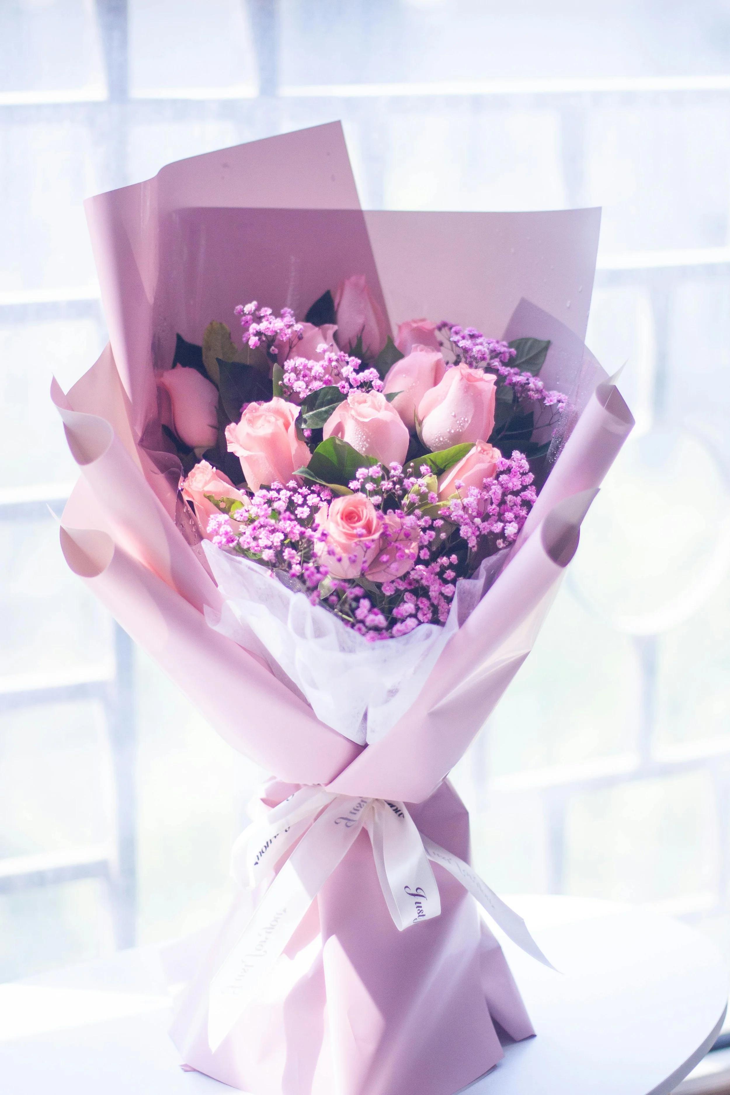 A bouquet of pink roses and purple baby's breath flowers wrapped in pink and purple paper with a white ribbon, set against a backlit window.