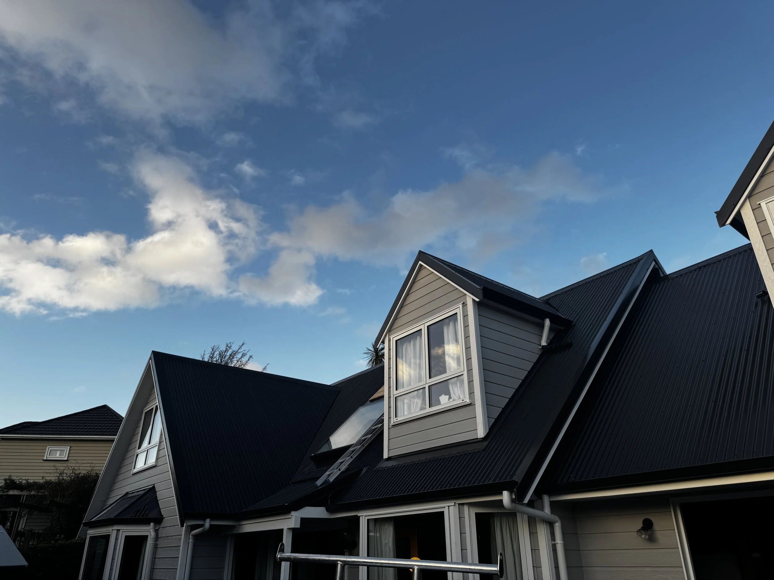 Photo of a house with dark metal roofing, dormer windows, and a partly cloudy sky overhead.