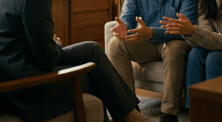 People in a therapy or counseling session, sitting on chairs and engaging in a discussion with hand gestures.
