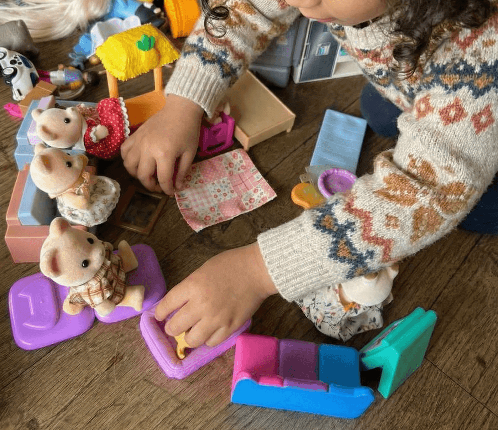 Child playing with various stuffed animals and toy furniture on a wooden floor.