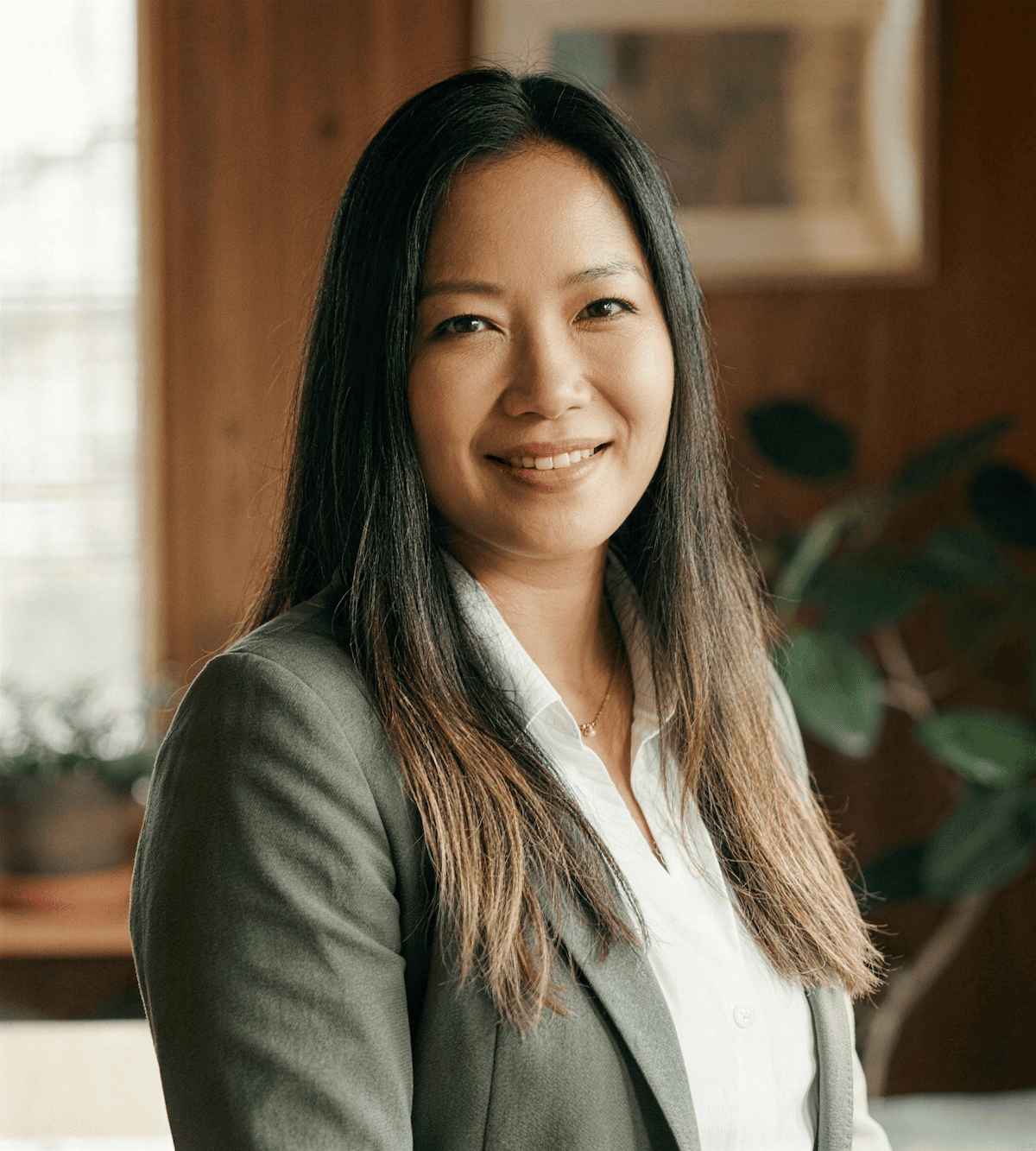 Portrait of a woman with long black hair, wearing a white blouse and gray blazer, smiling indoors with a plant and wooden wall background.