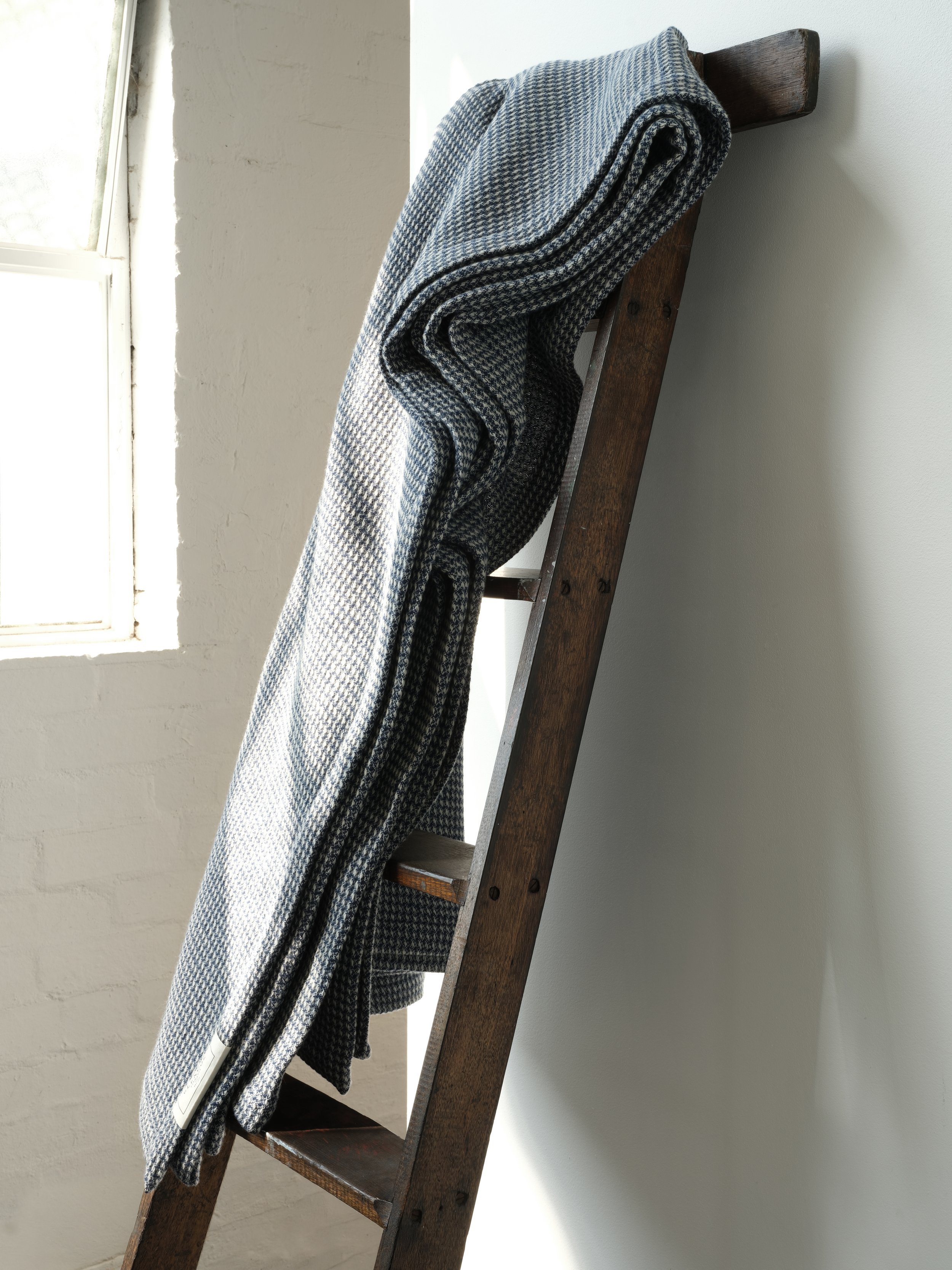 A ladder leaning against a white wall with a window nearby. A gray and black patterned blanket is draped over one of the ladder's rungs.