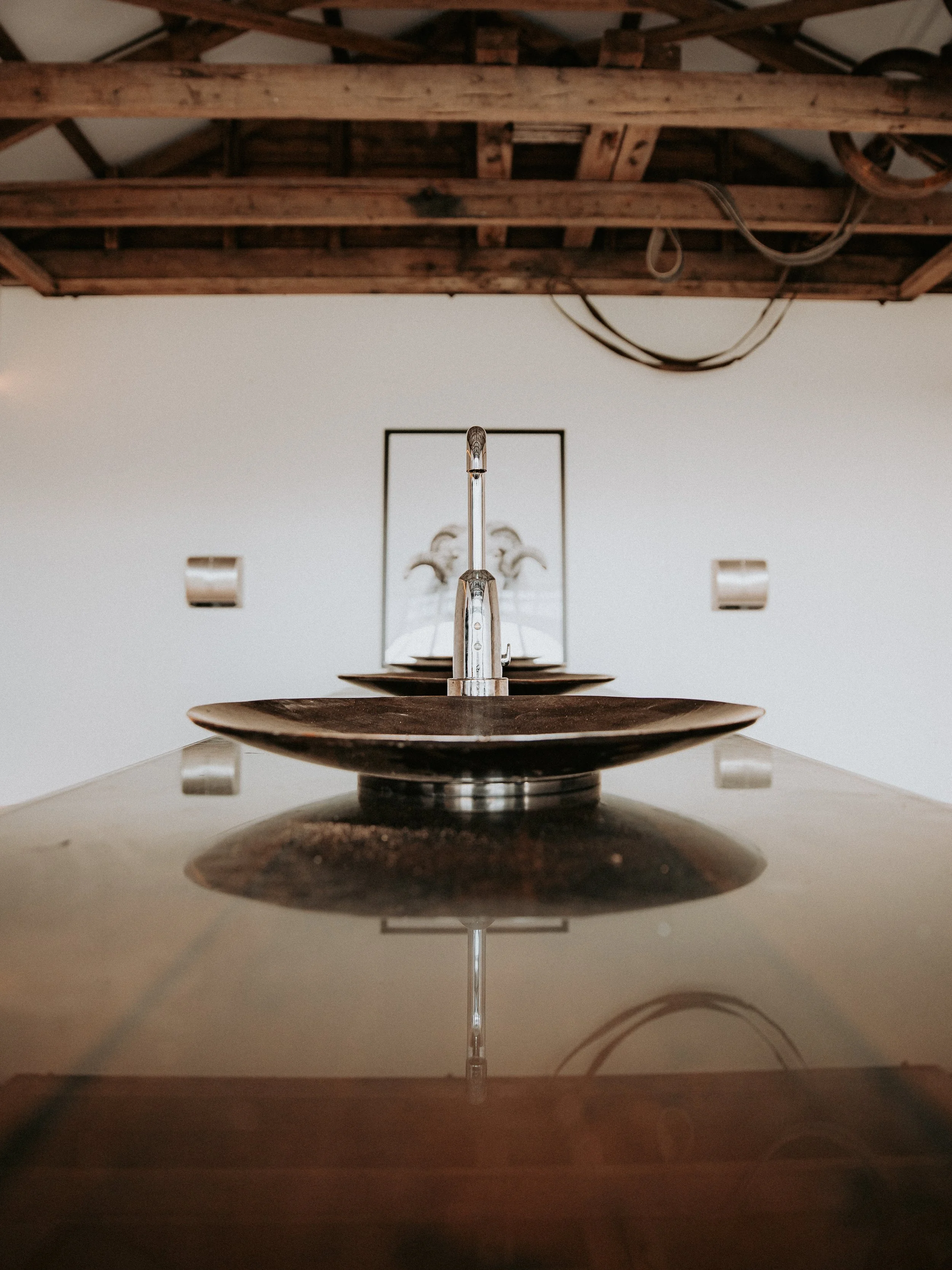 A photo taken from below showing a wooden ceiling with beams, a metal faucet with a reflection, and a mirror with a chandelier reflected in it.