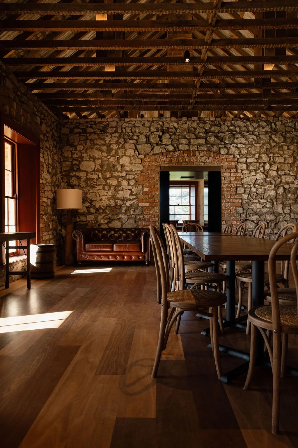 Interior of a rustic dining room with exposed stone and brick walls, wooden ceiling beams, a large wooden table surrounded by wooden chairs, and a brown leather sofa with a floor lamp nearby.