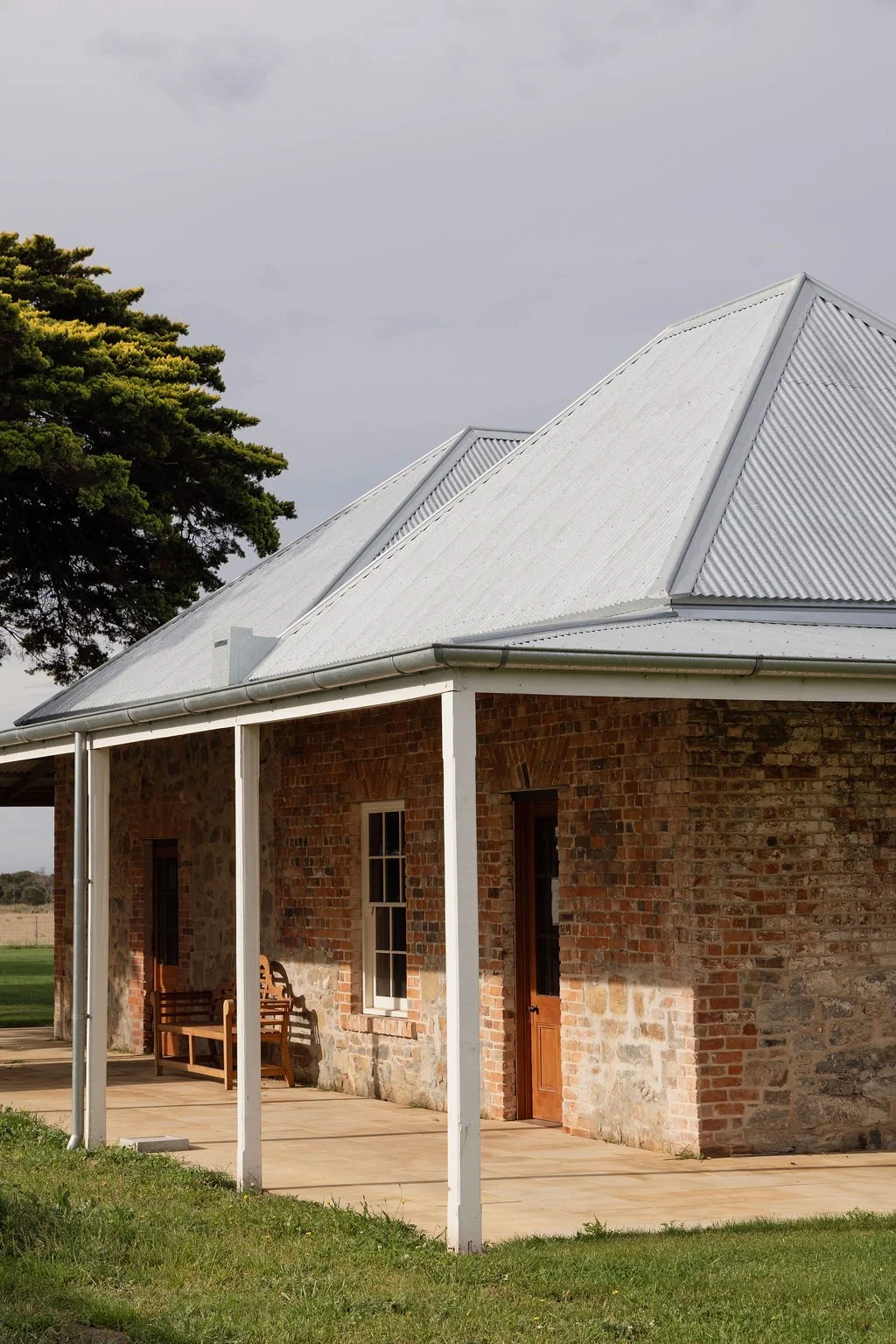Brick building with a metal roof, a porch with benches, and a large tree in the background under a cloudy sky.