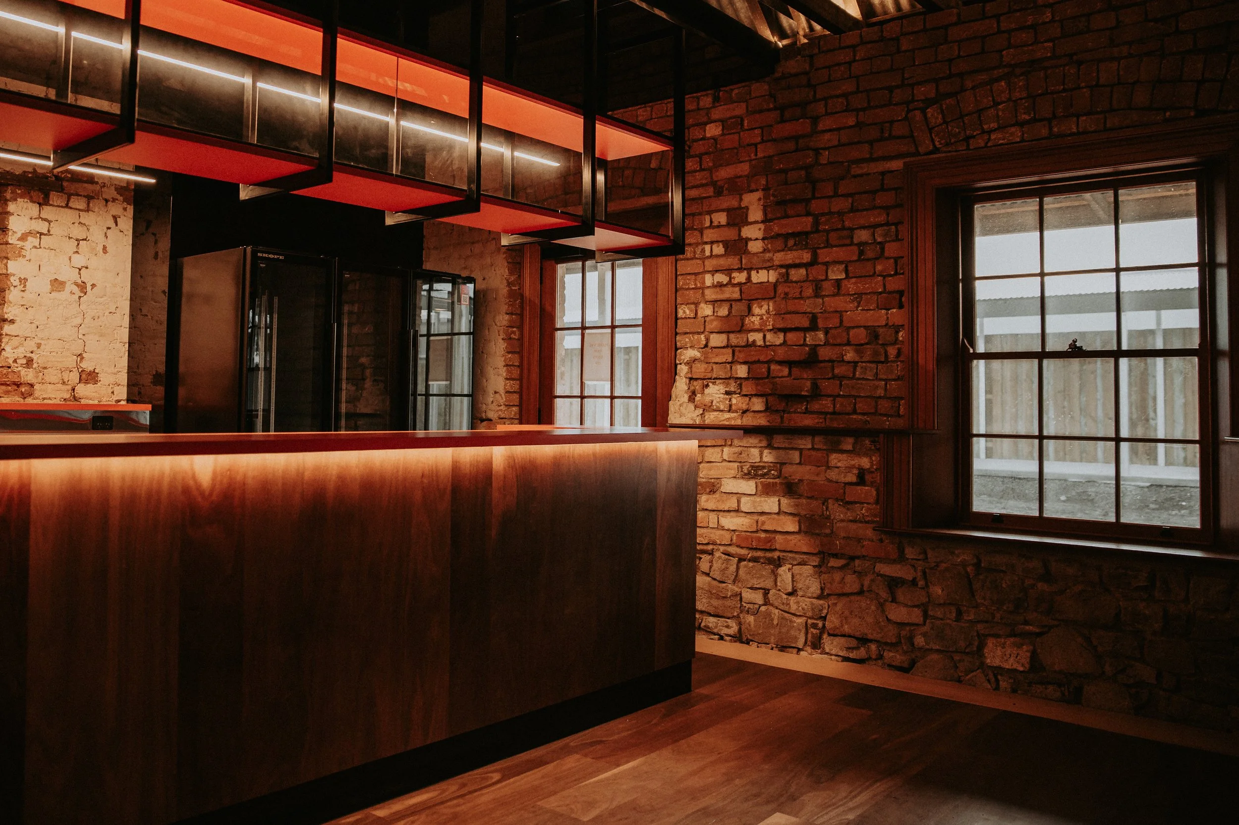 Interior of a room featuring exposed brick walls, large windows with black frames, a wooden counter, and a modern hanging light fixture with red and black accents.