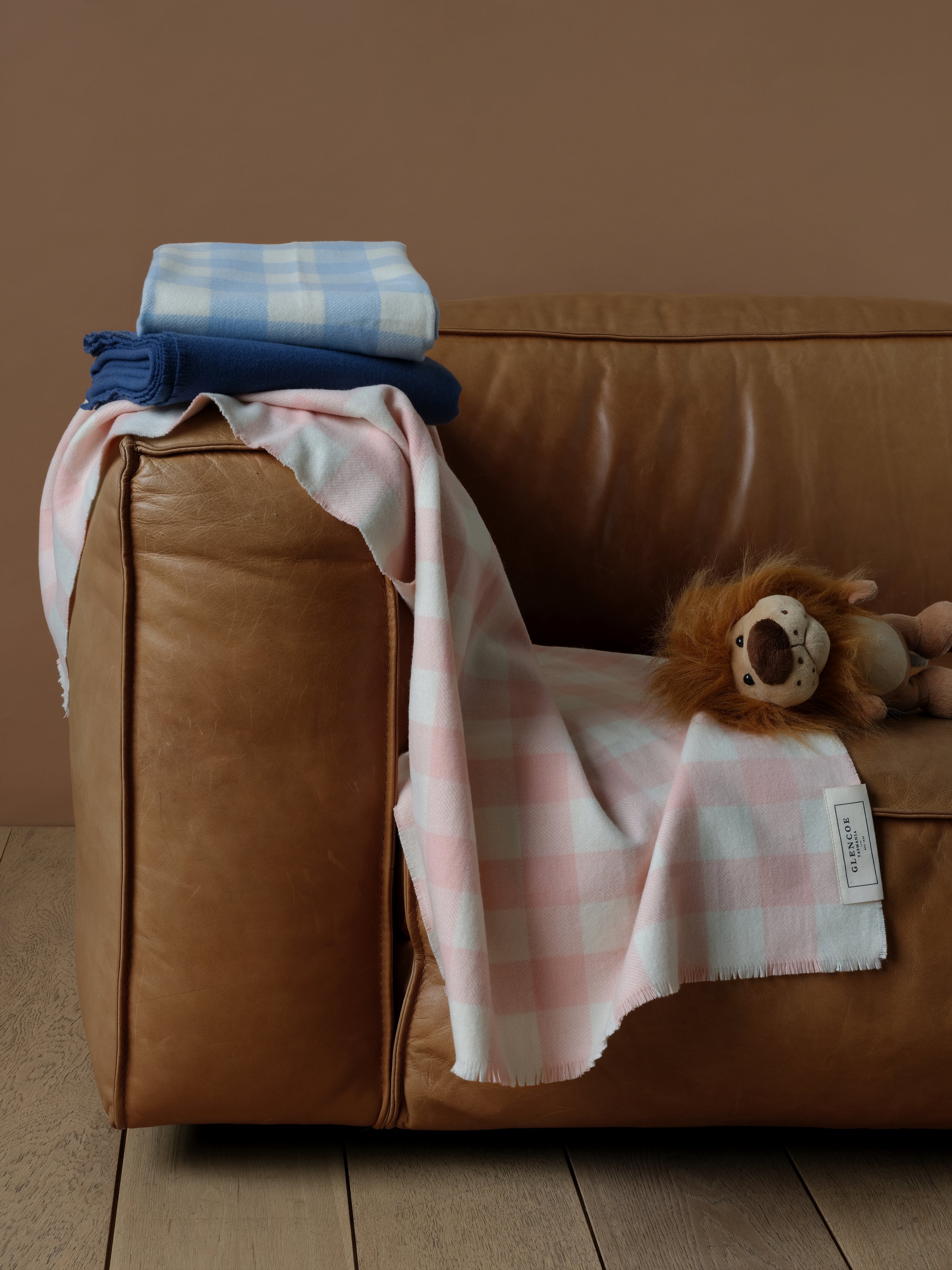 A brown leather lounge chair with a blanket, a folded checkered cloth, and a stuffed lion toy placed on it. The chair is next to a wall and on a tile floor.