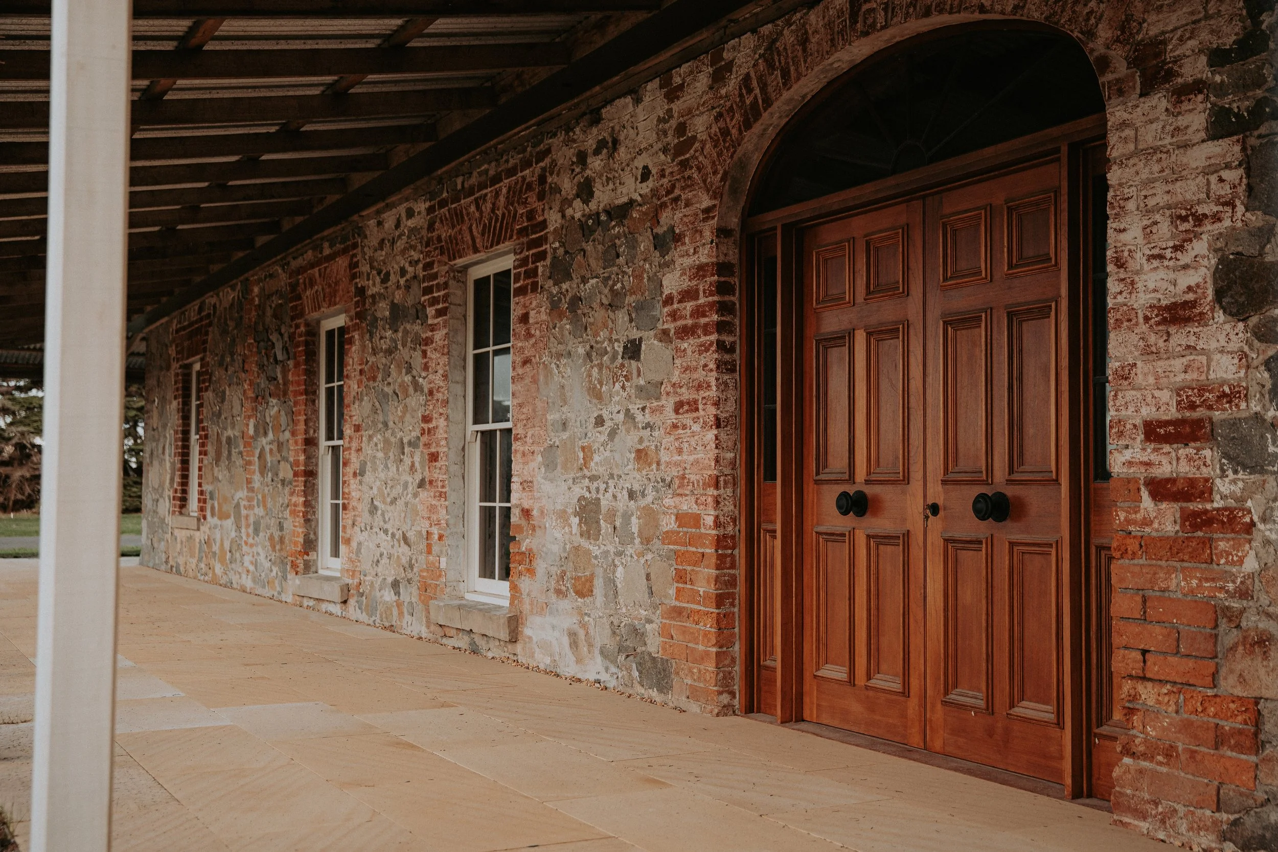 Close-up of a stone and brick house exterior with a large wooden front door, two windows, and a porch area.
