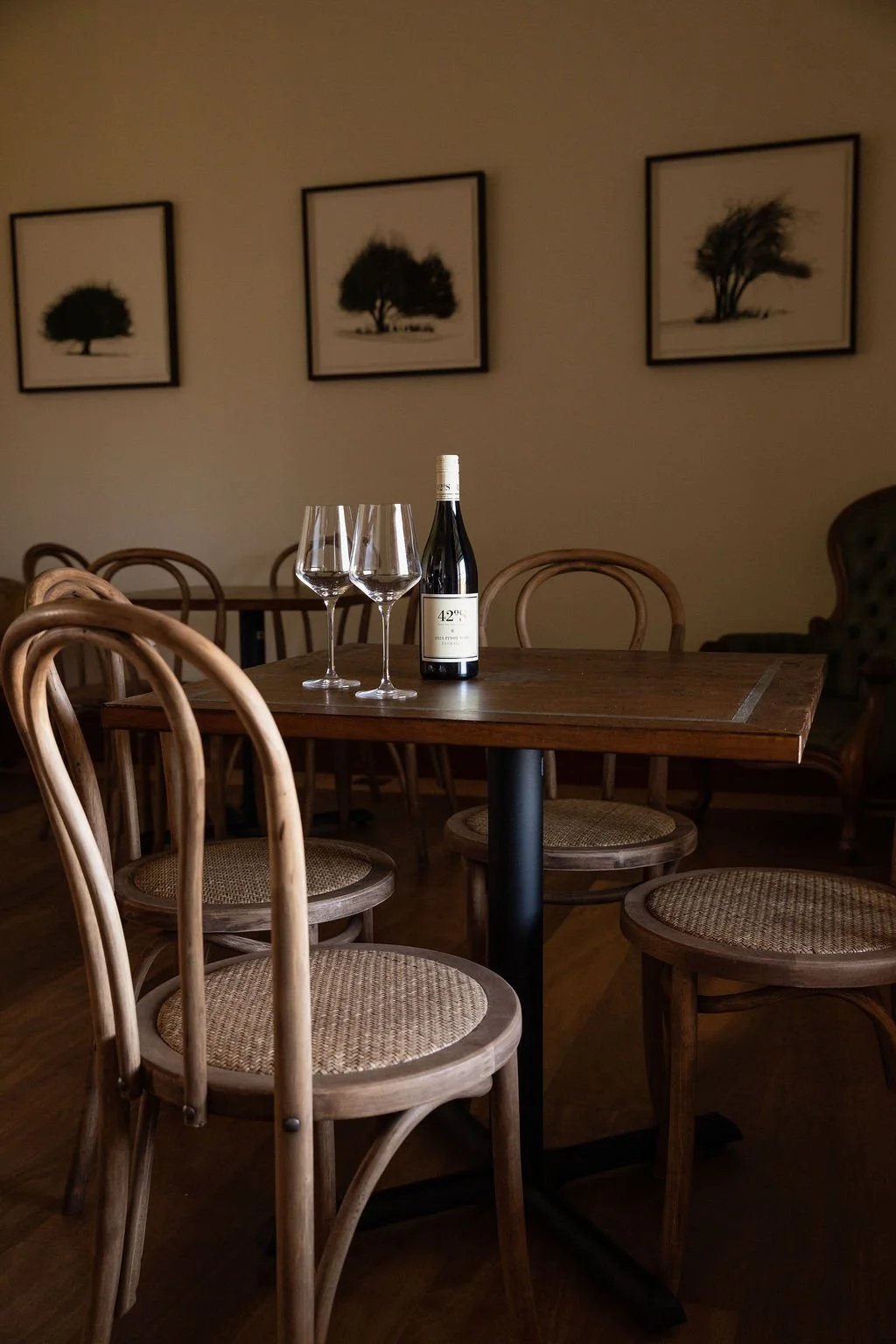 Empty wooden dining table with two wine glasses and a bottle of wine, surrounded by wooden chairs, in a dimly lit room with framed black-and-white tree photographs on the wall.