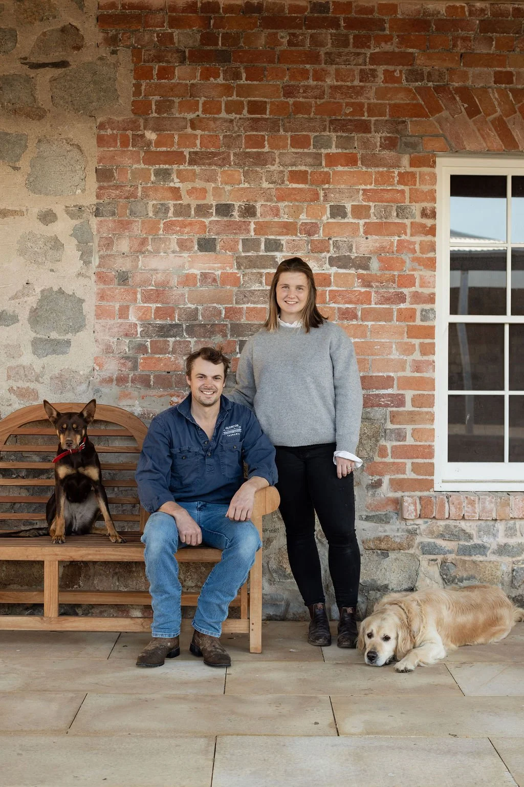 A man and a woman with two dogs pose in front of a rustic brick wall. The man is sitting on a wooden bench with a black and brown dog beside him, and the woman is standing next to him, smiling. The woman has shoulder-length brown hair and is wearing a gray sweater and black pants. The dog on the bench is sitting upright, while the other dog lying on the floor is a golden retriever, resting with its head on the ground.