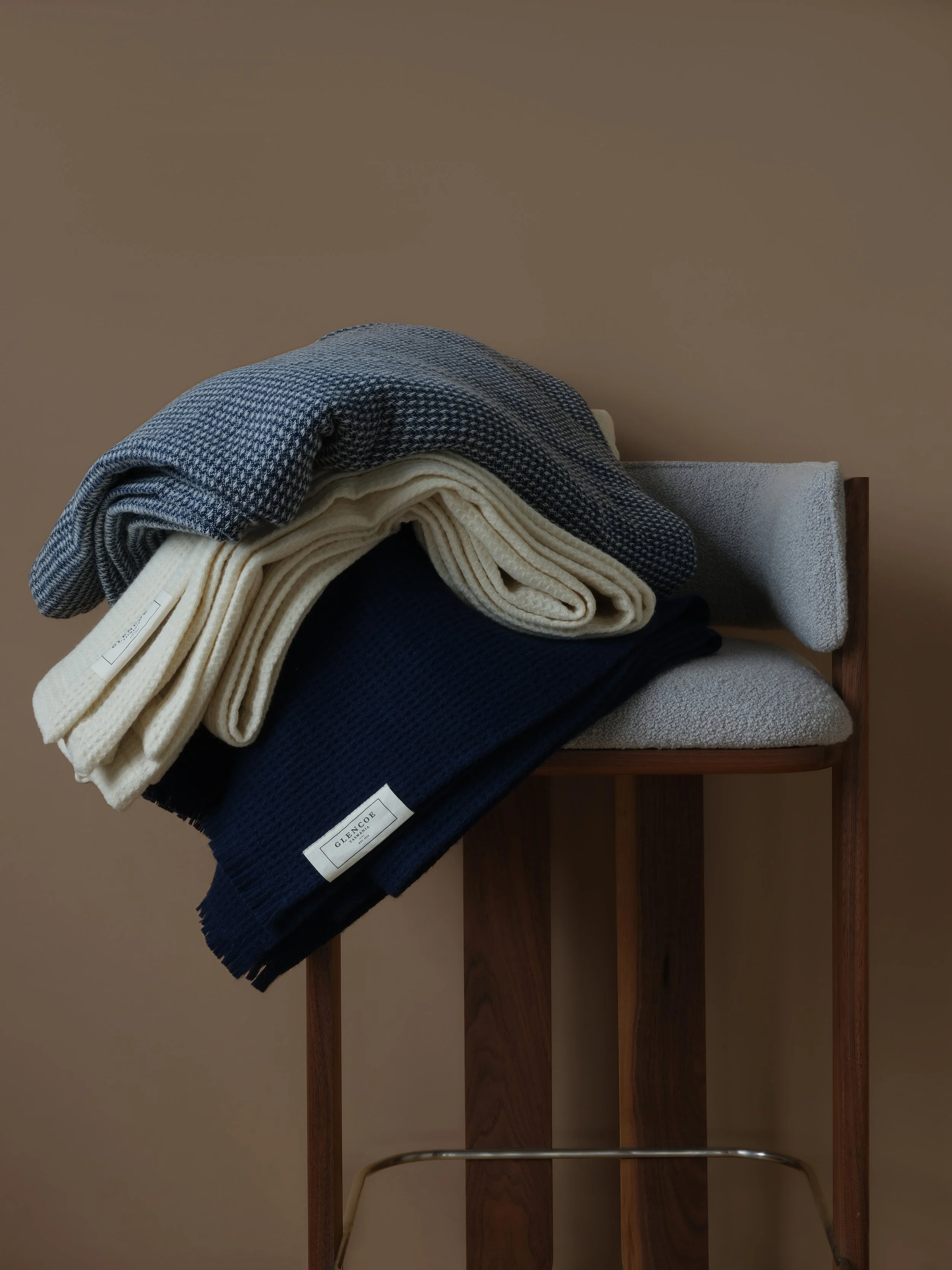 A wooden rack with folded clothing, including a checkered pattern shirt, cream-colored fabric, navy fabric, and grey textured fabric, against a beige wall.