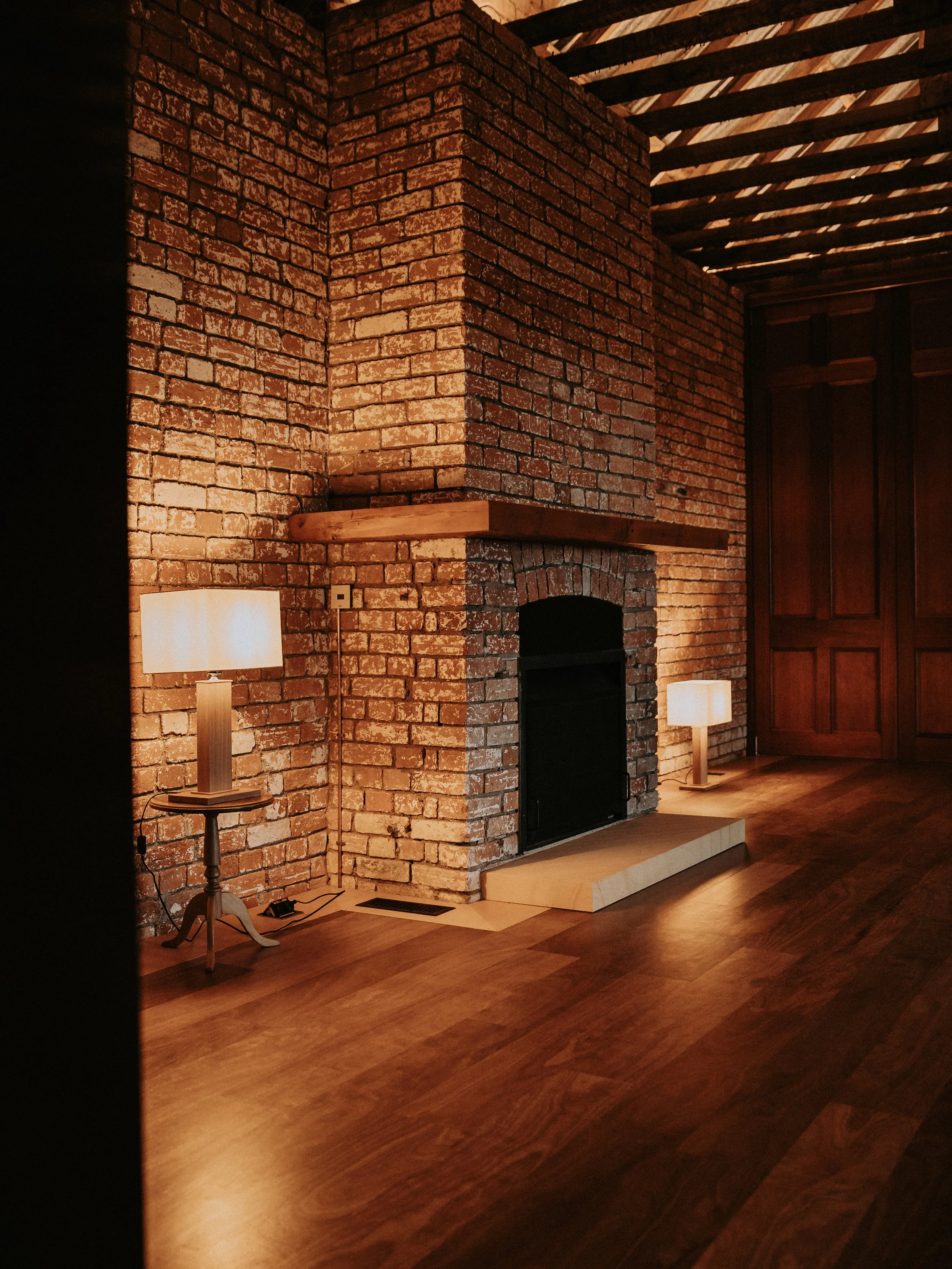 Brick fireplace with a wooden mantel, two lamps with beige shades on either side, and a wood-paneled wall to the right. Hardwood floor.
