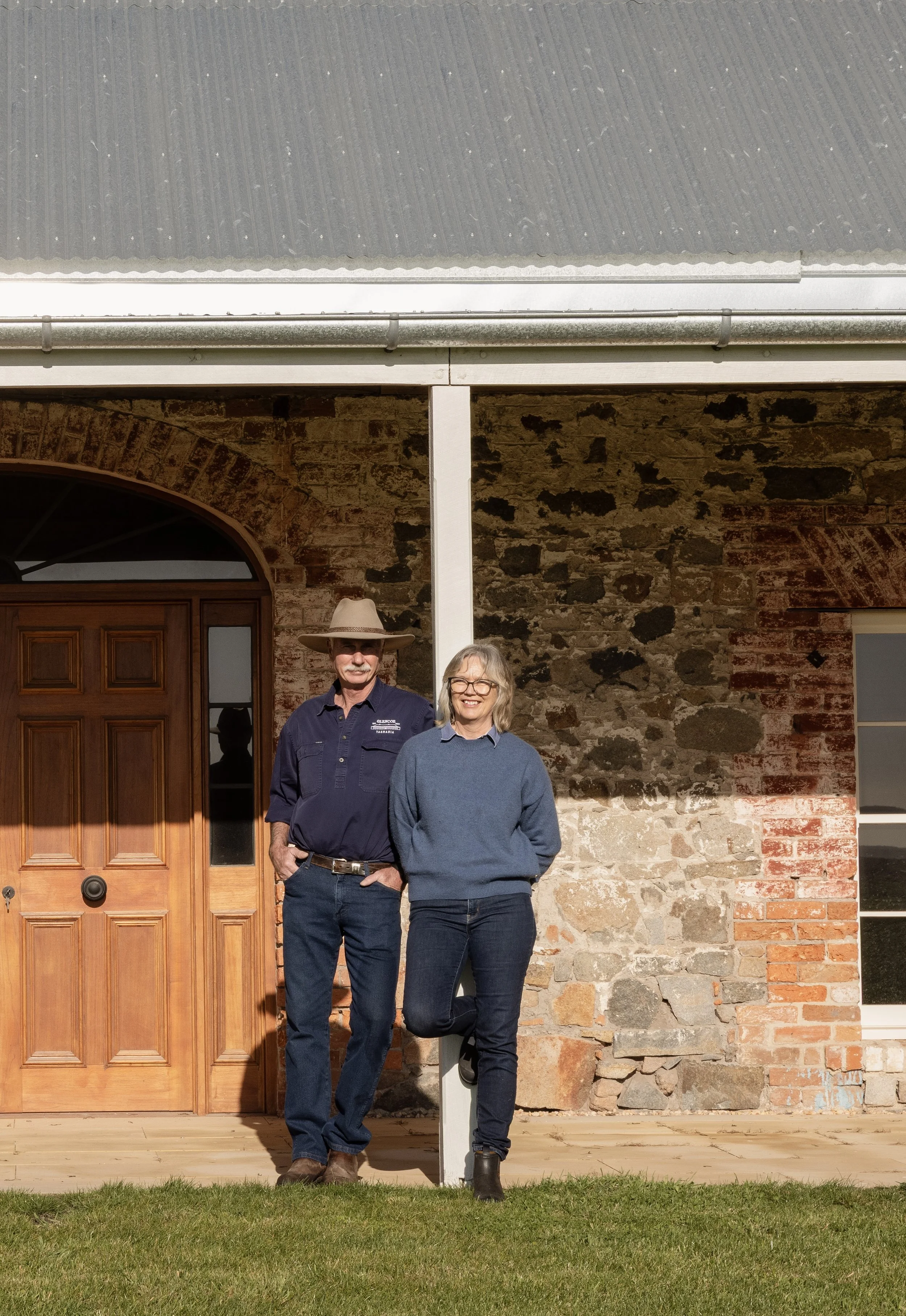 A man and a woman standing in front of a rustic brick house with a wooden front door, a window, and a gray metal roof. They are smiling and posing for the photo, with the woman wearing glasses and a blue sweater, and the man wearing a hat and a navy blue shirt.