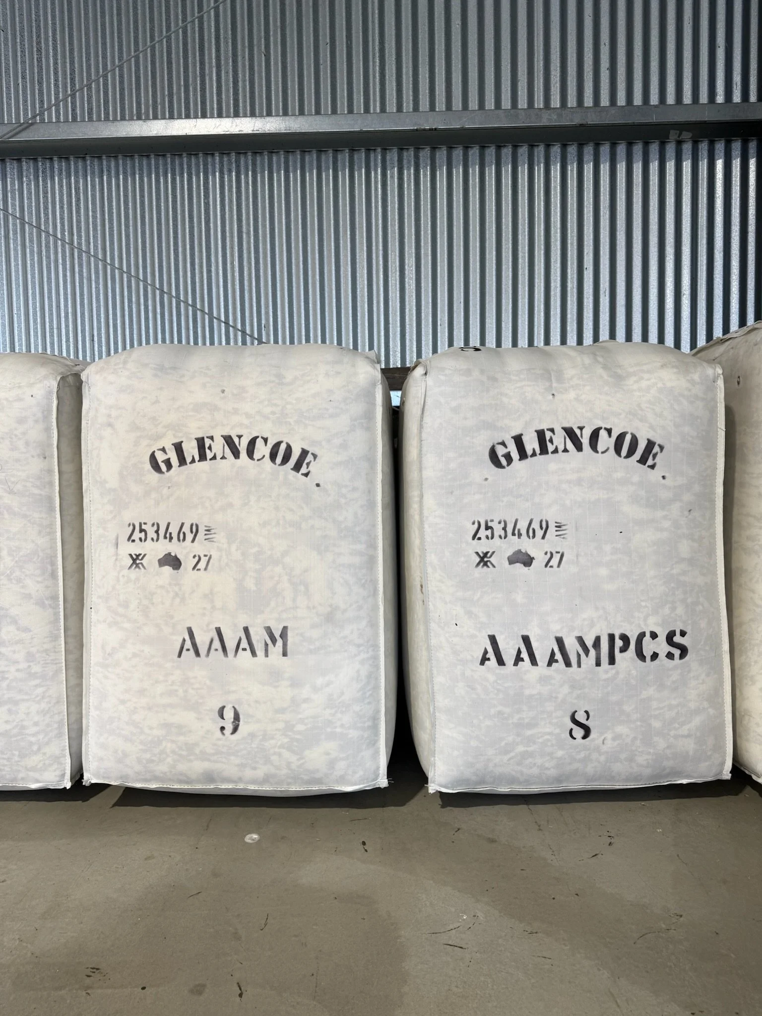 Stacked agricultural bags labeled 'Glencoe' with text 'AAAM' and 'S' on them, in an industrial setting with metal wall background.