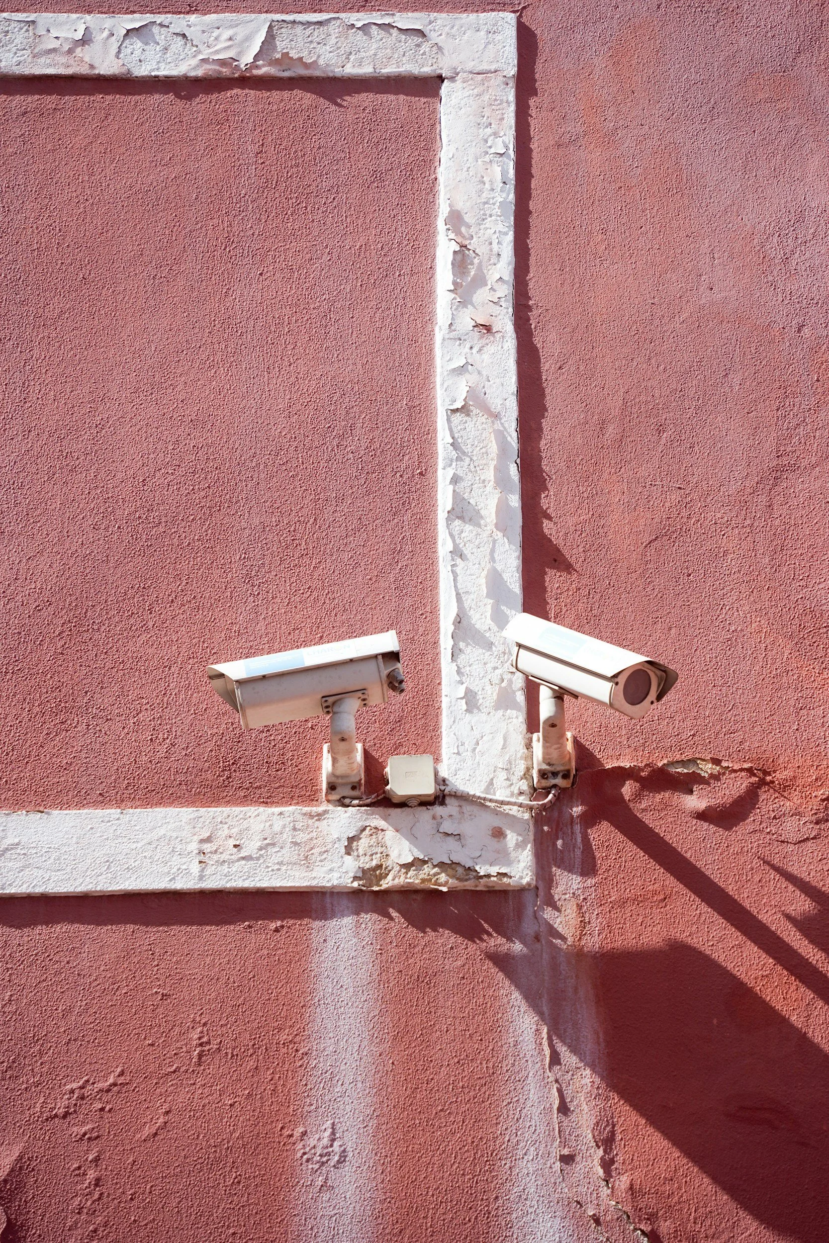 Two security cameras mounted on a pink stucco wall with peeling white trim and a visible vertical crack.