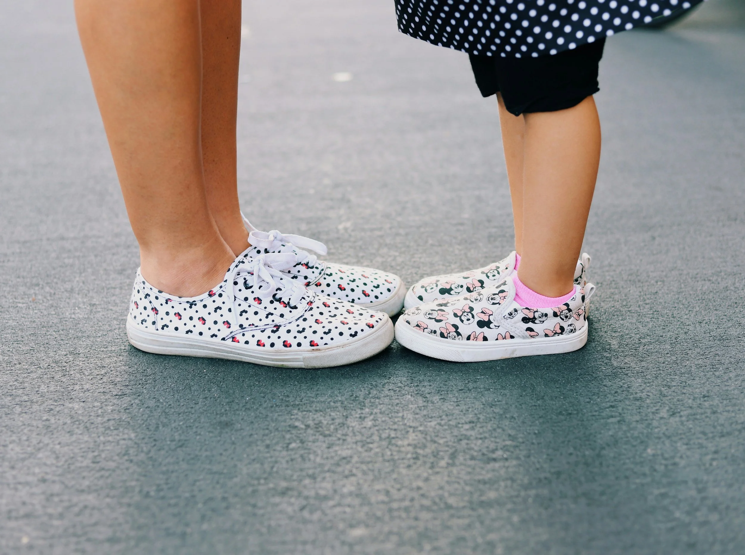 Close-up of two pairs of children's feet wearing Minnie Mouse themed sneakers standing on a paved surface.