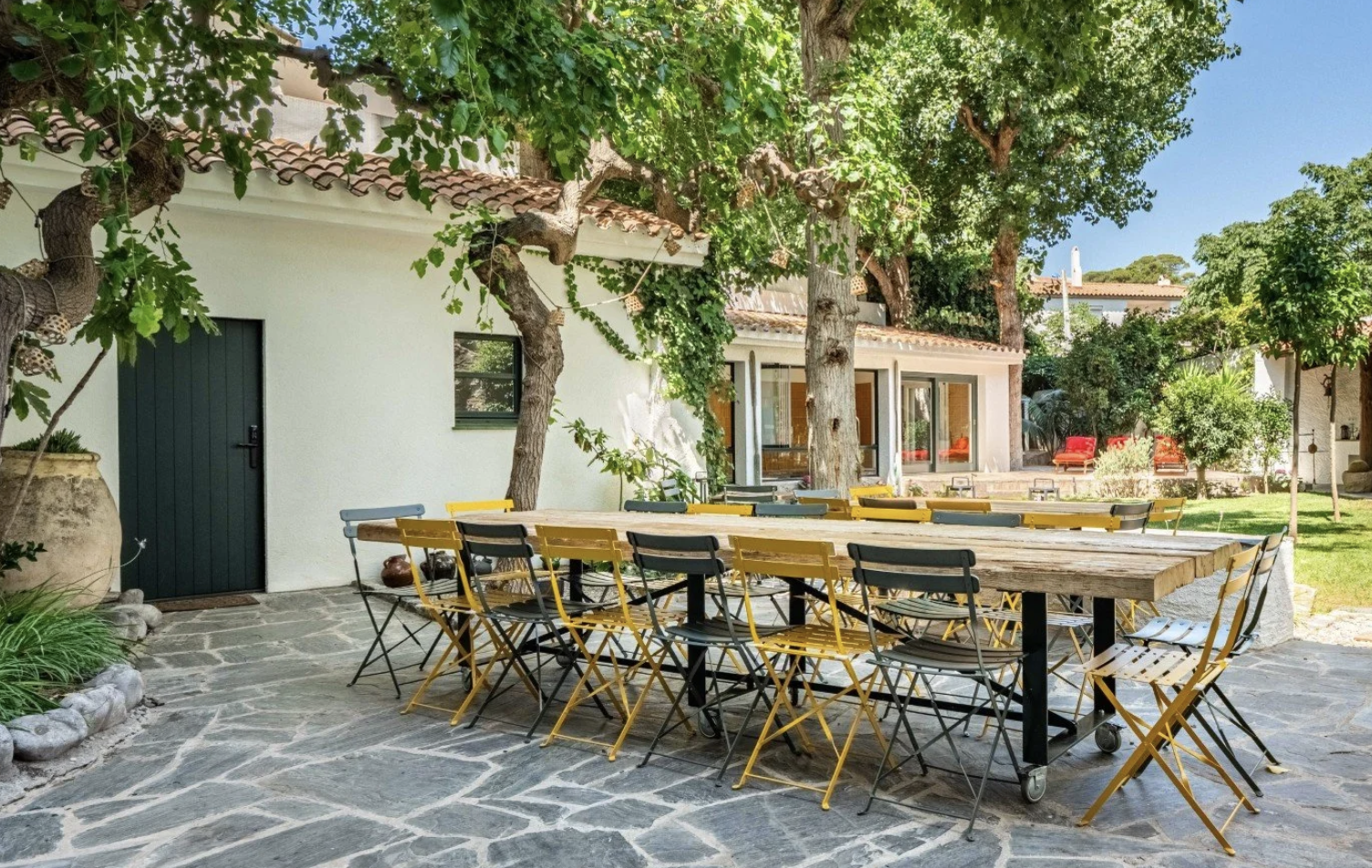 Garten mit langer Esstischgruppe, umgeben von Bäumen, vor weißem Haus mit Terrassen und Stühlen, sonnig und im Freien
