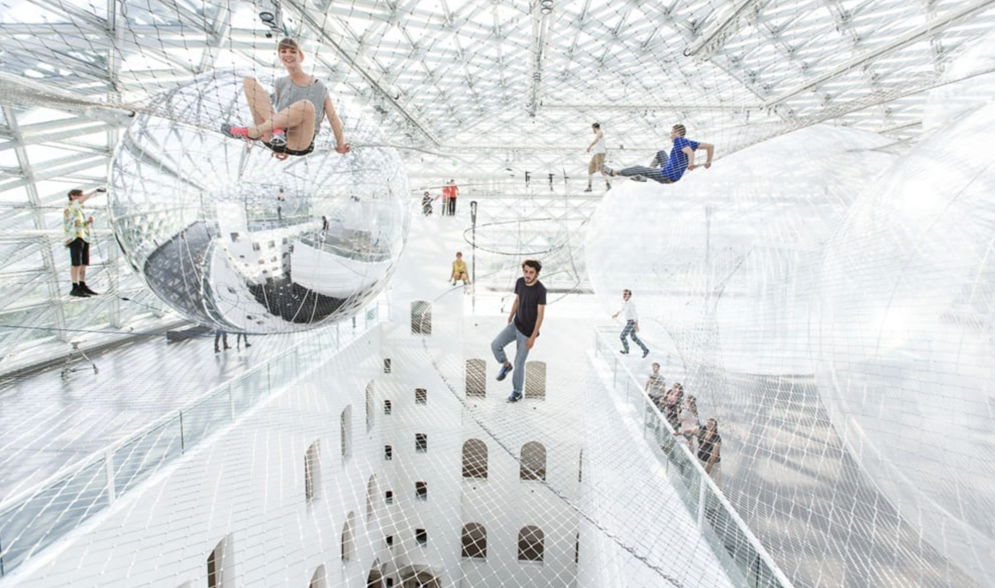 Menschen in einer futuristischen, transparenten Spielhalle mit Netzen, Spiegelkugeln und Kletterelementen.