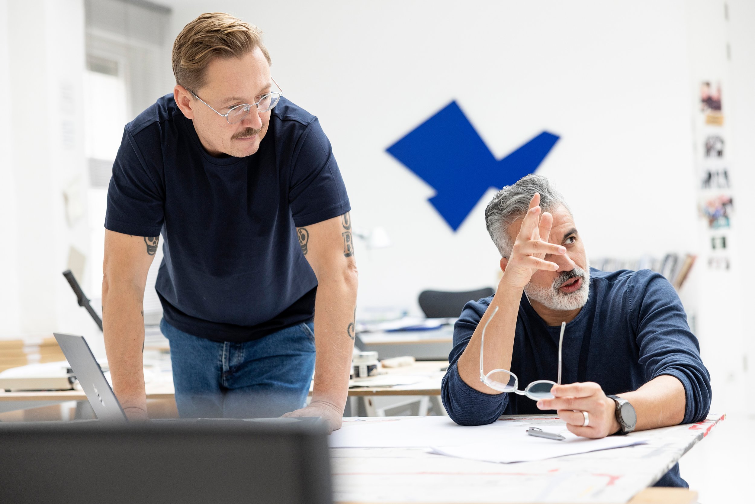 Zwei Männer in einem Büro, der eine steht, der andere sitzt und zeigt auf die Wand. Im Hintergrund eine große blaue Pfeilmarkierung an der Wand.