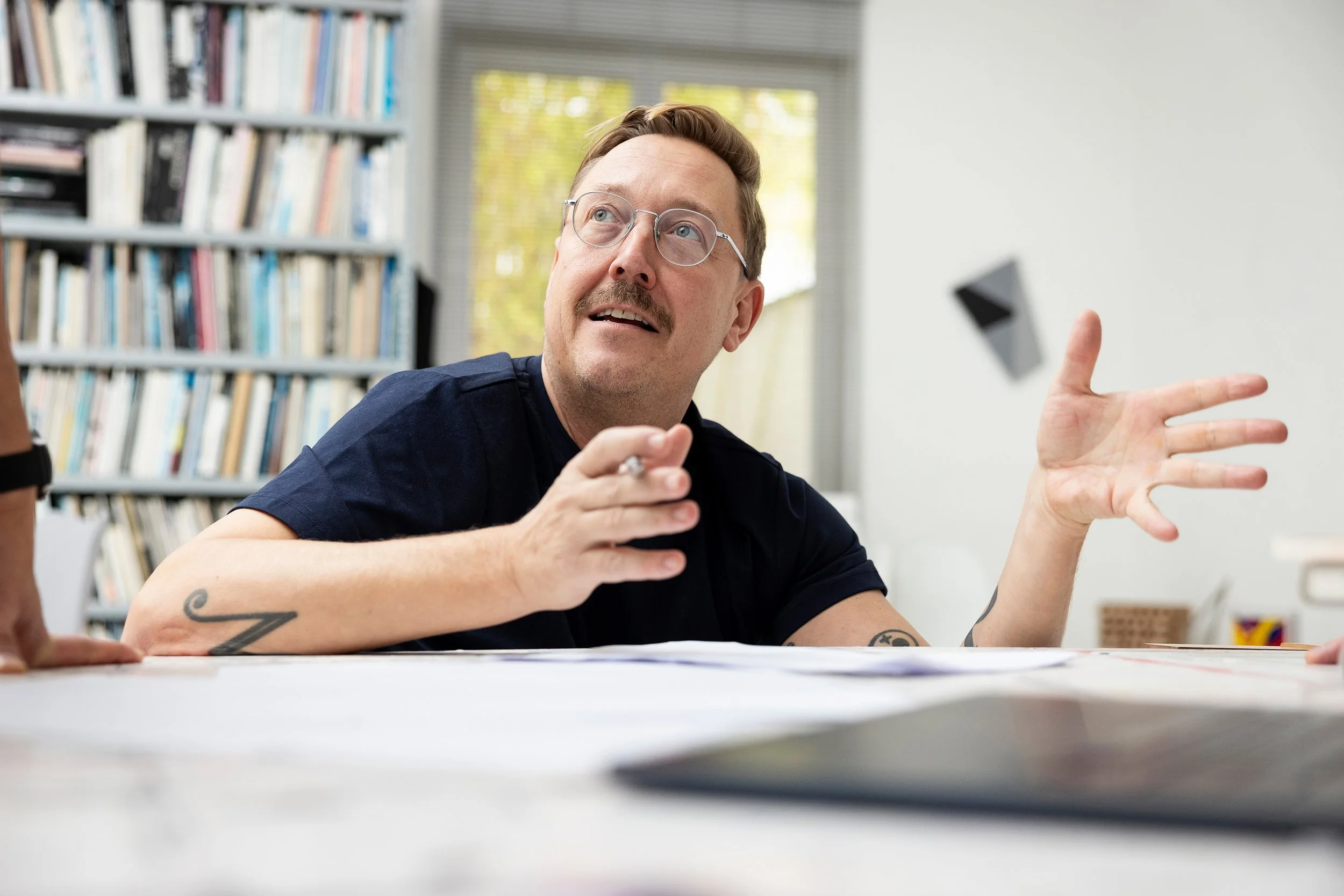 Mann mit Brille und Tätowierungen, spricht in einem Büro mit Bücherregal im Hintergrund, verwendet Handgesten während eines Gesprächs.