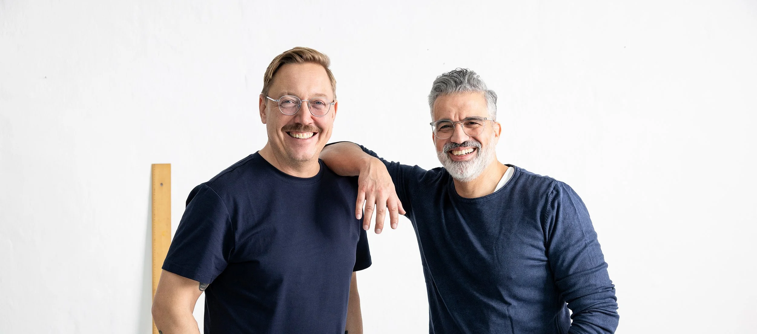 Zwei lachende Männer in blauen T-Shirts posieren vor weißer Wand, einer legt Arm auf Schulter des anderen.