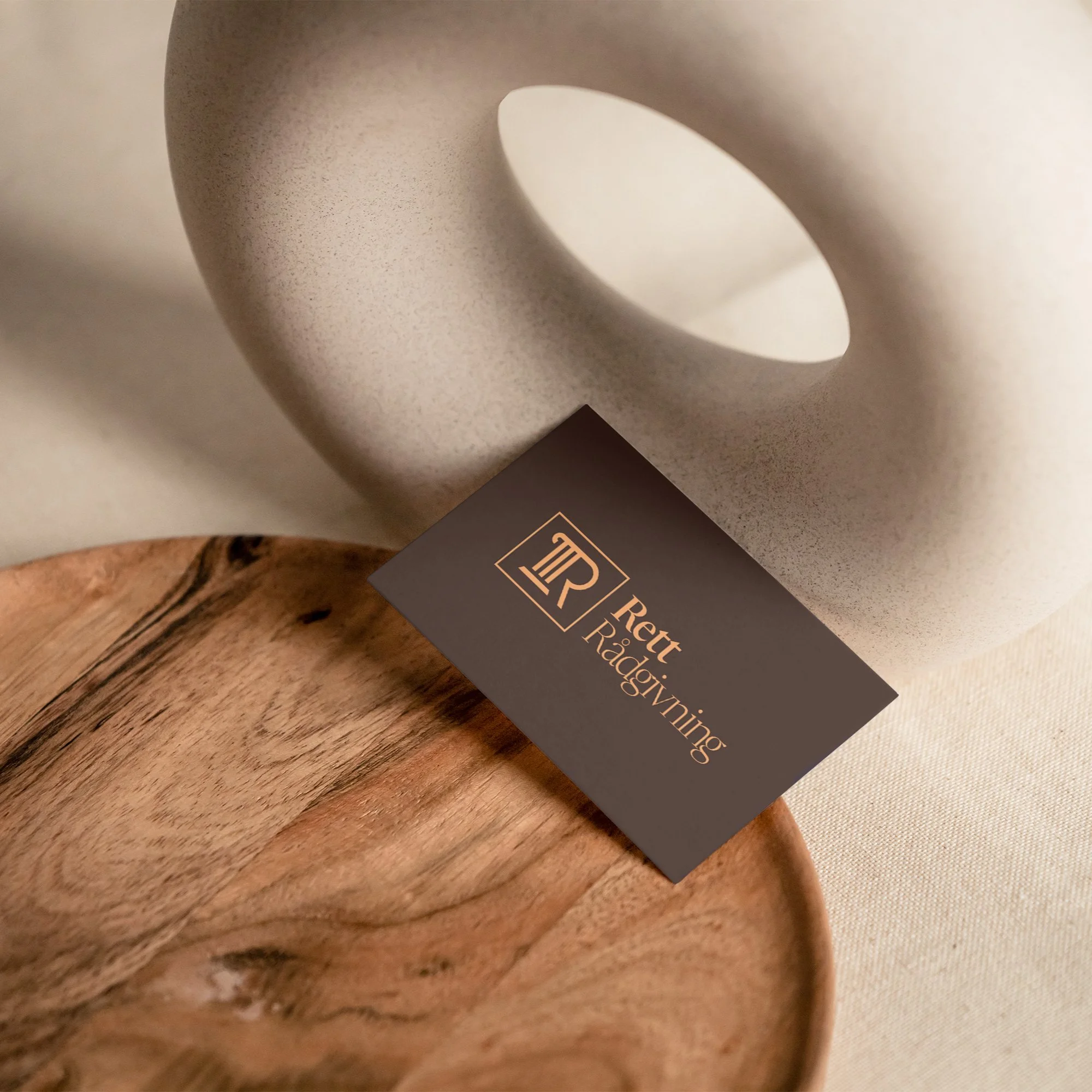 Et firkantet kort med logo og tekst, plassert på en trefat, foran en lys, rund, hullformet keramikkdekorasjon.