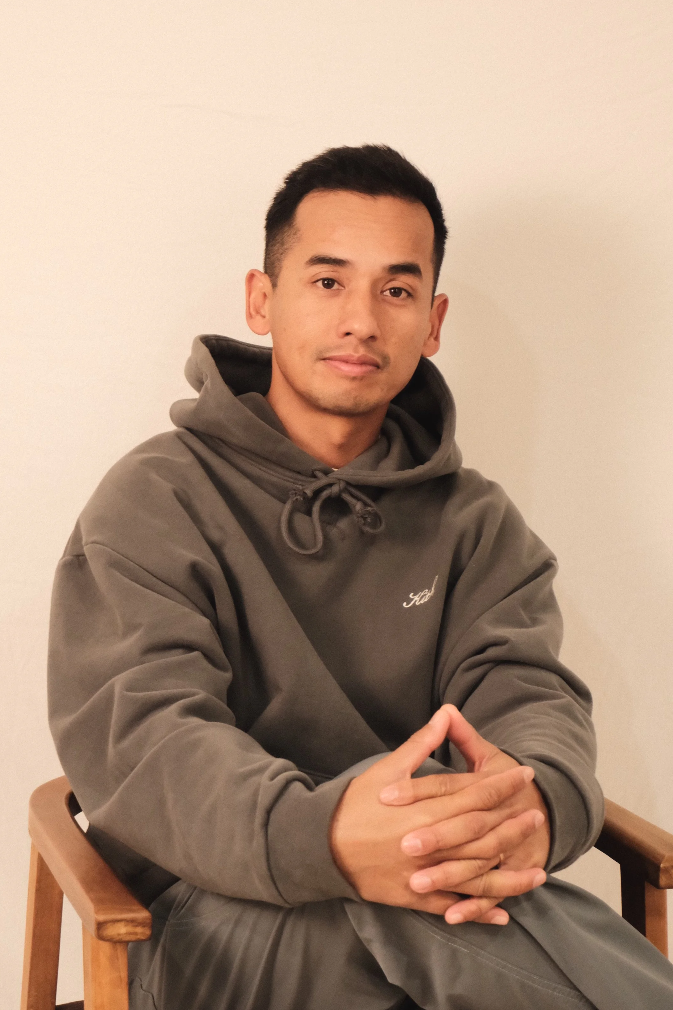 A young man standing indoors in front of glass doors, wearing a white hoodie, a black puffer vest, a white cap, and khaki pants.