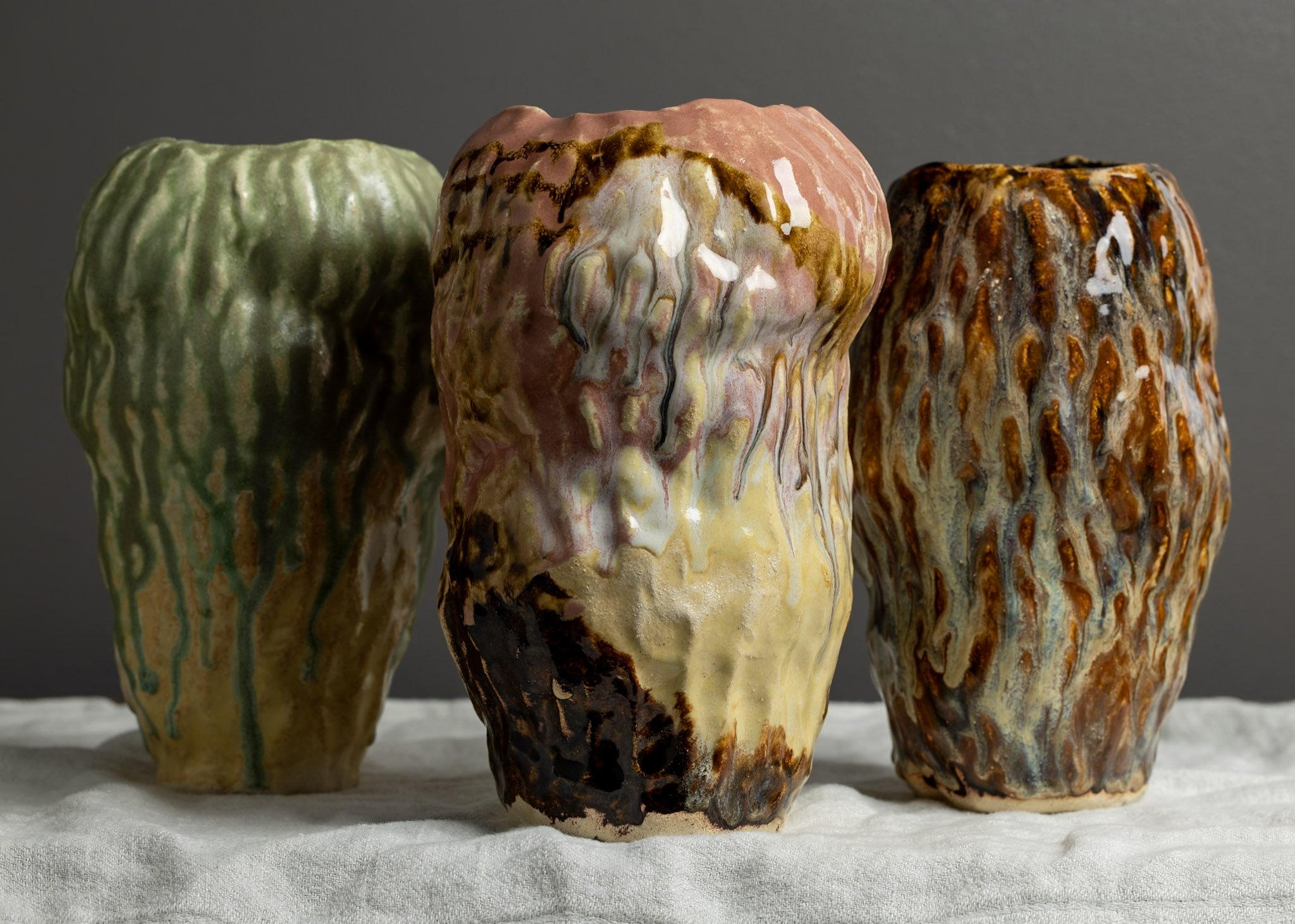 Three glazed ceramic vases with drippy, colorful glaze patterns placed on a white surface against a gray background.