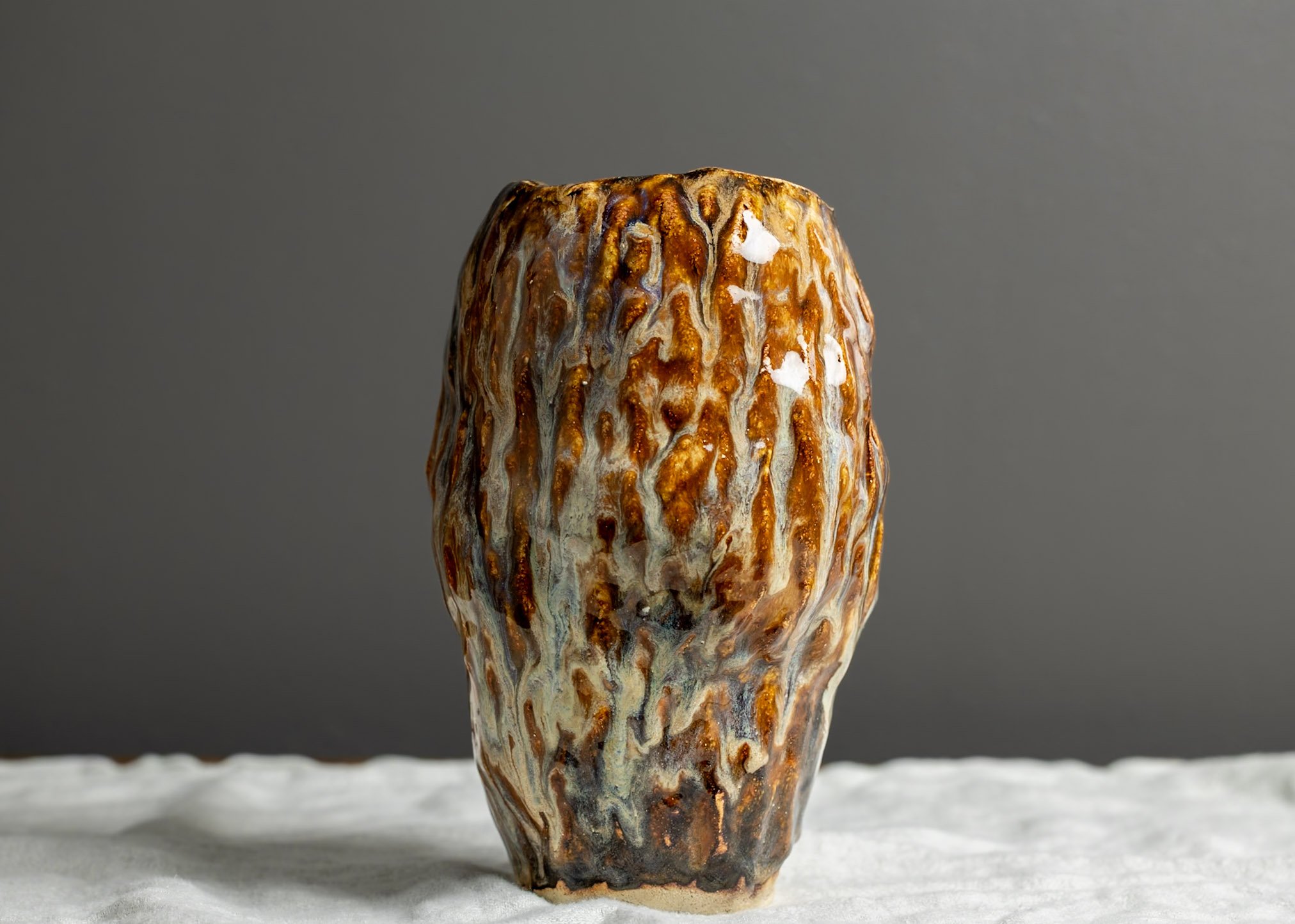 Close-up of a ceramic vase with a textured, multicolored glaze, placed on a white surface against a gray background.