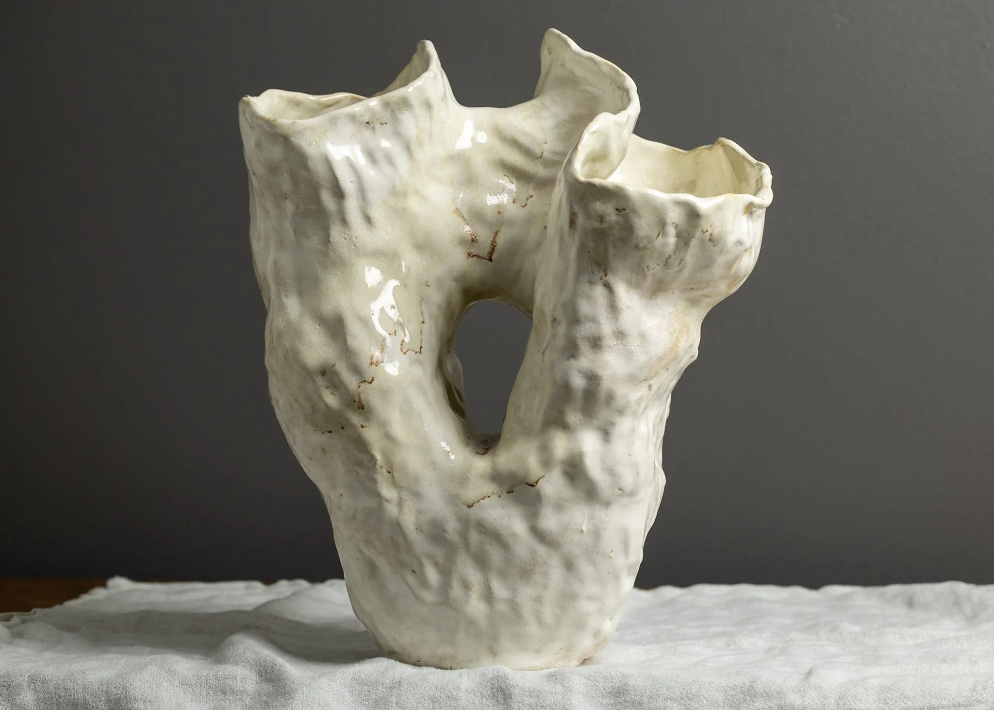 A ceramic sculpture resembling a tree or branch with a textured surface, glazed in an off-white color with some brownish accents, placed on a white surface with a gray background.