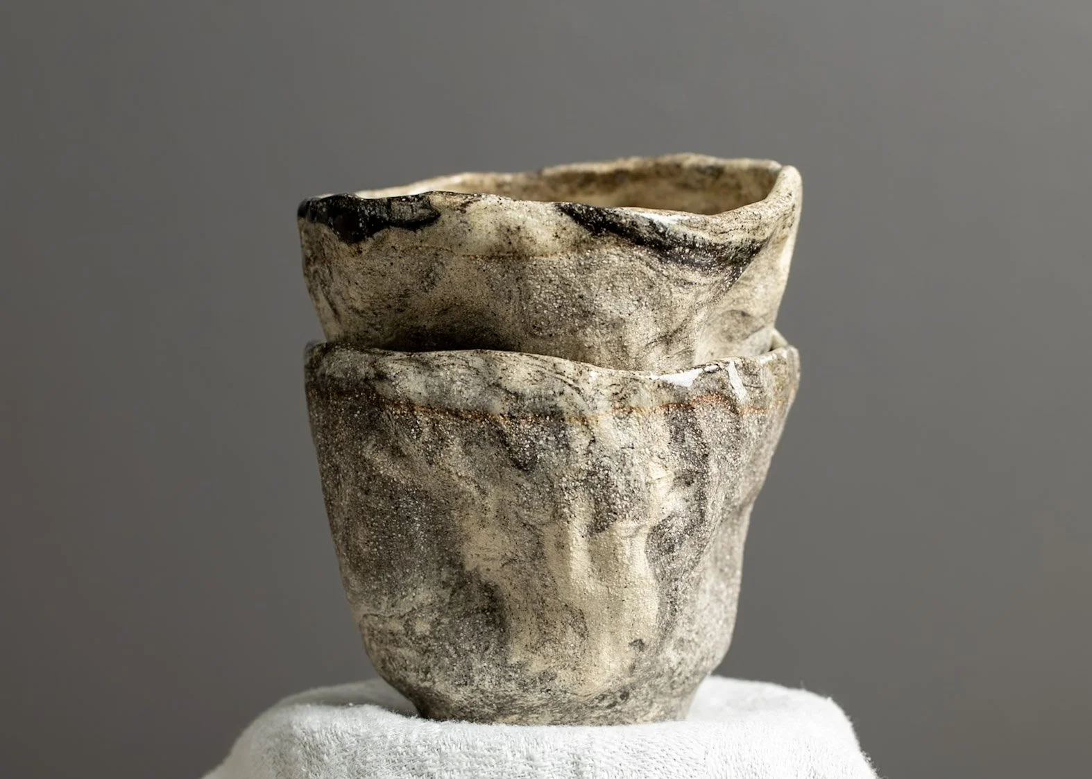 A ceramic vessel with an uneven, textured surface and earthy colors, stacked on top of a similar vessel, set against a neutral gray background.