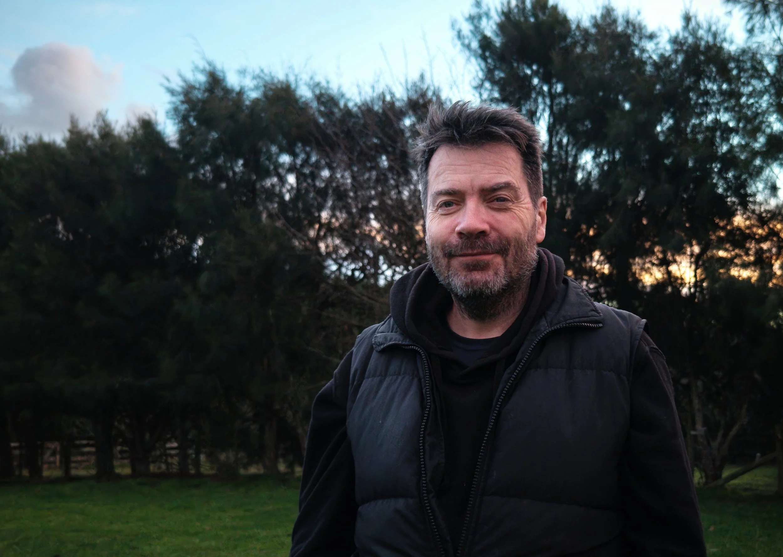 Man with dark hair and beard wearing a black puffy vest standing outdoors with trees and a cloudy sky in the background.