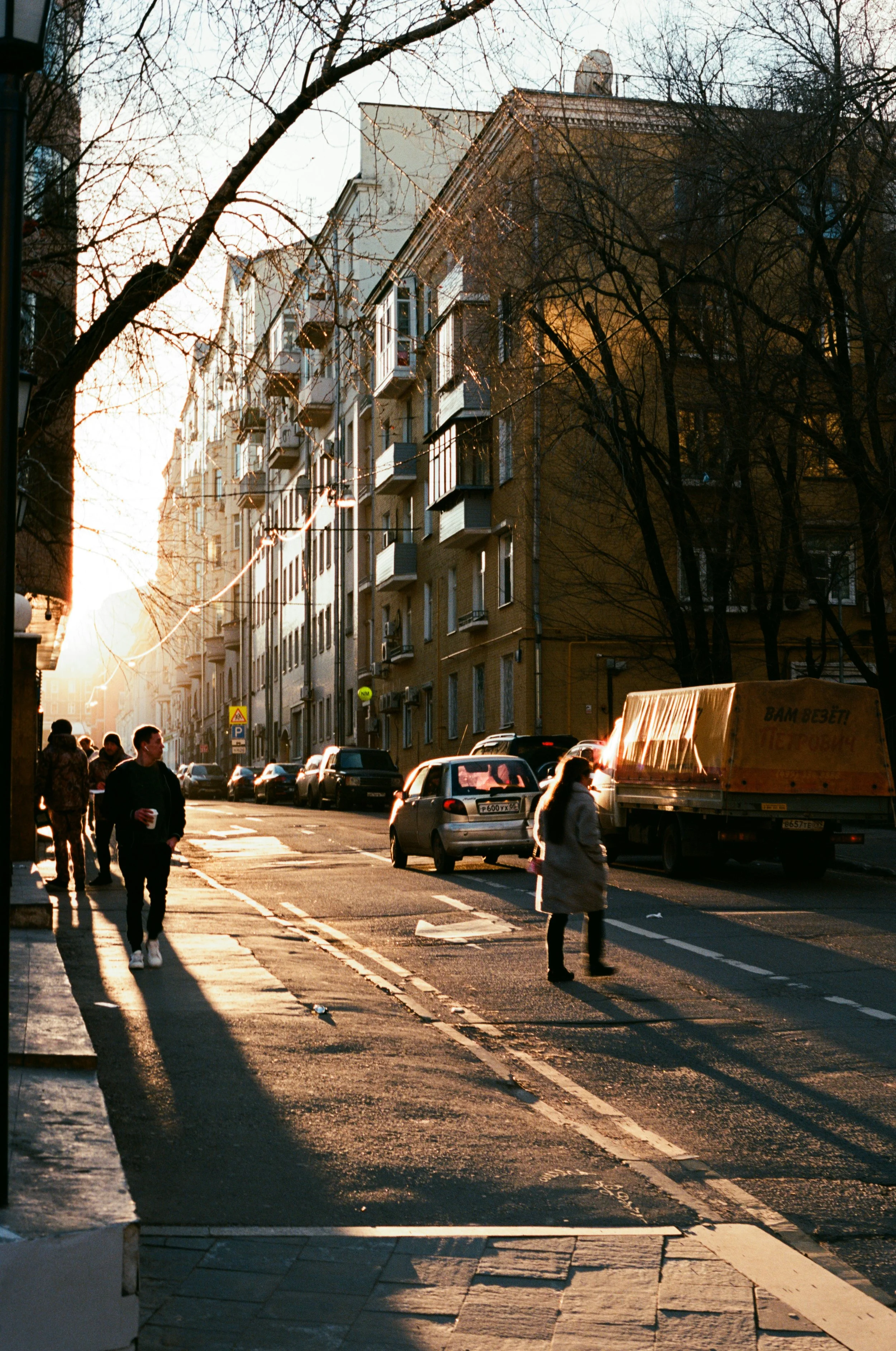 A city street at sunset with pedestrians walking on the sidewalk and cars parked along the side. Buildings with balconies line the street, and leafless trees cast shadows. The scene is illuminated by warm sunlight.