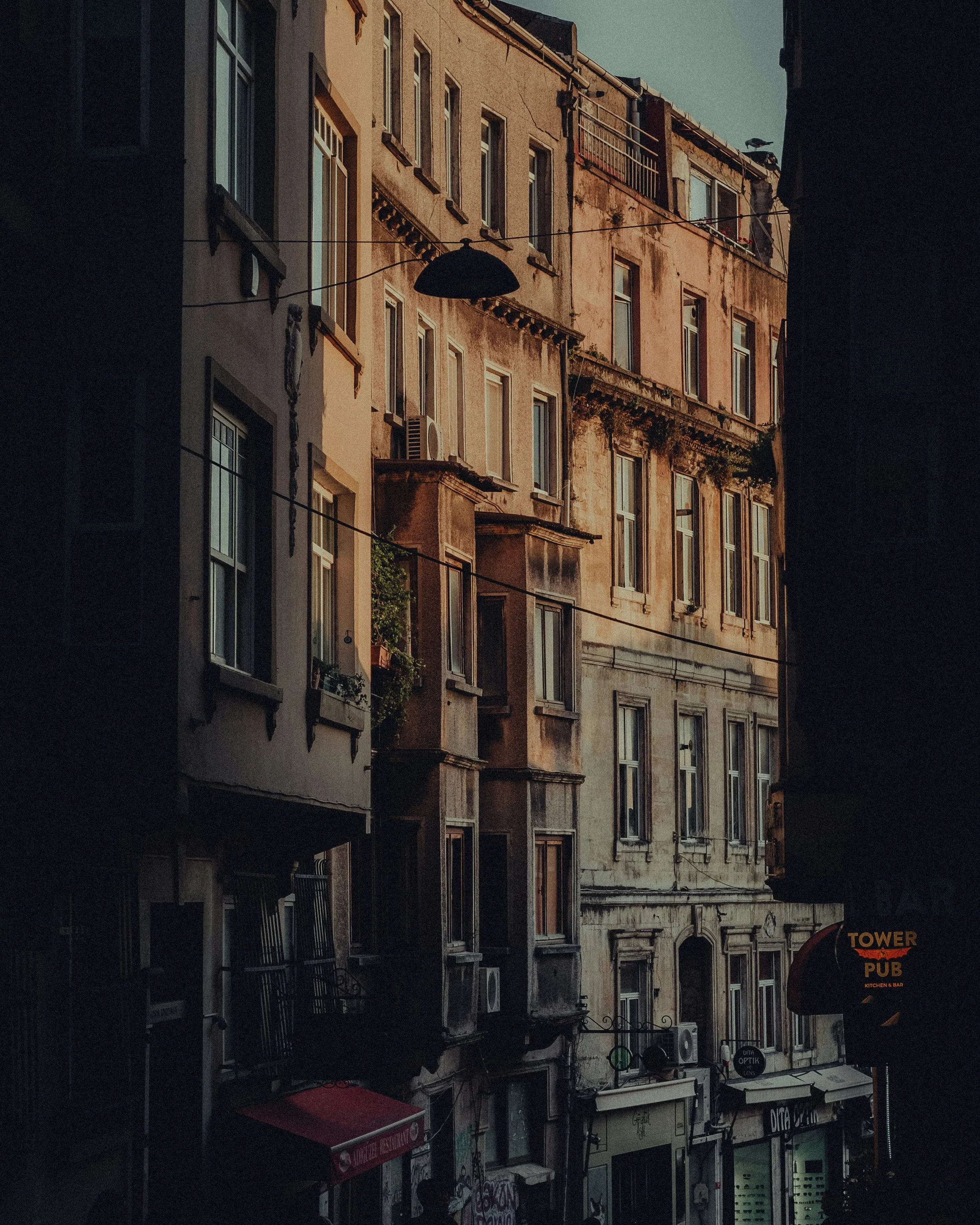 Old multi-story buildings with worn facades along a narrow street, late afternoon sunlight casting shadows, with signs for a pub and various shops at the street level.