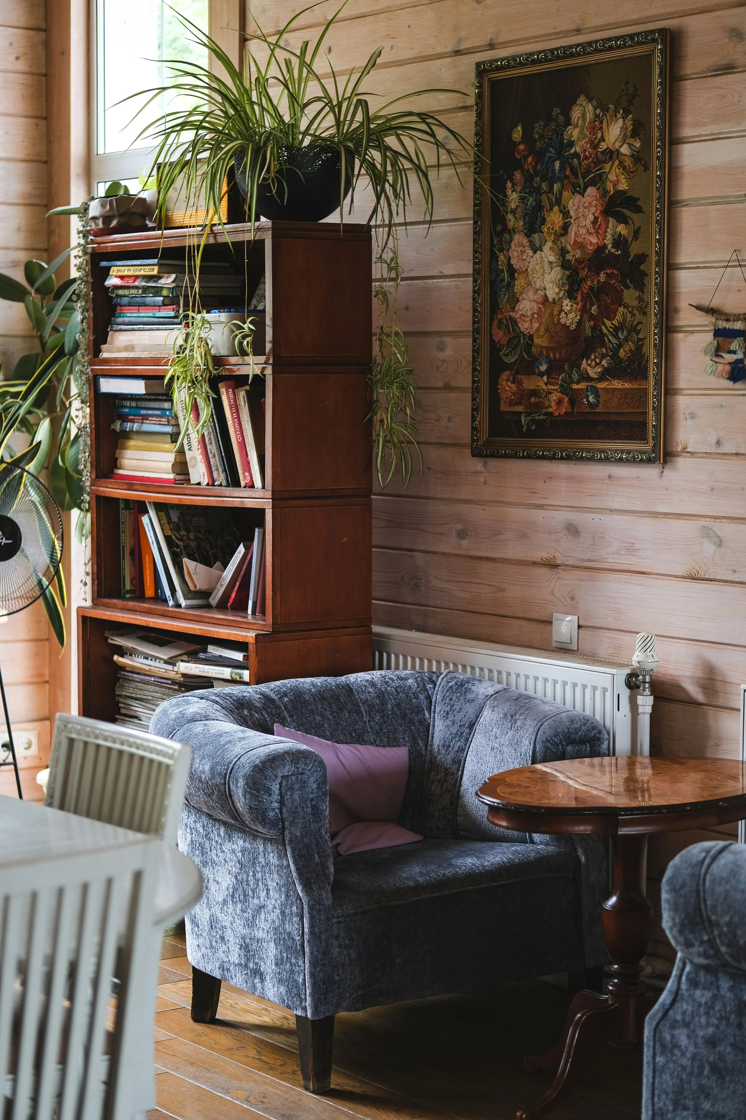Cozy living room with wooden walls, a blue velvet armchair with a purple pillow, a round wooden table, a bookshelf filled with books, a large potted plant on top of the bookshelf, and artwork of flowers on the wall.