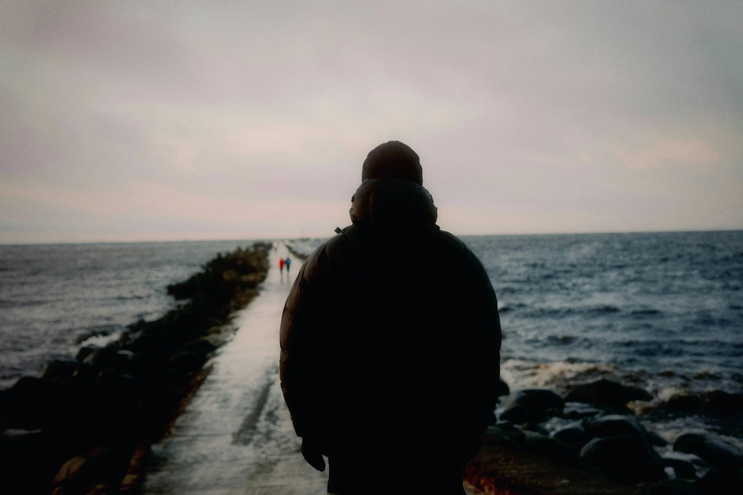 Person walking on a rocky pier by the ocean during cloudy weather, with other people in the distance.