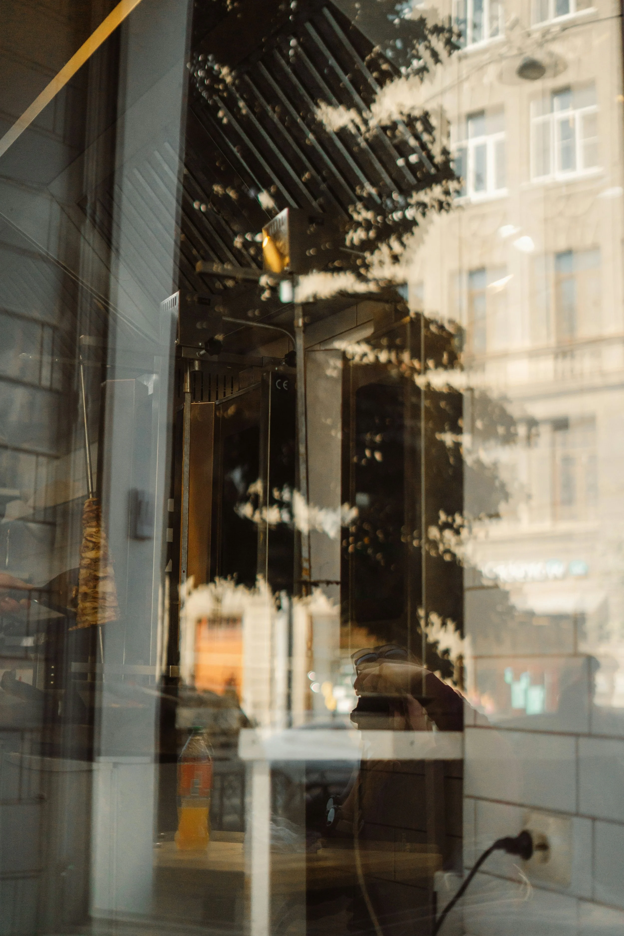 Reflection of city buildings and trees in a glass window of a restaurant or cafe; inside the cafe, a table with a bottle and electrical outlet, and a reflection of the person taking the photo.