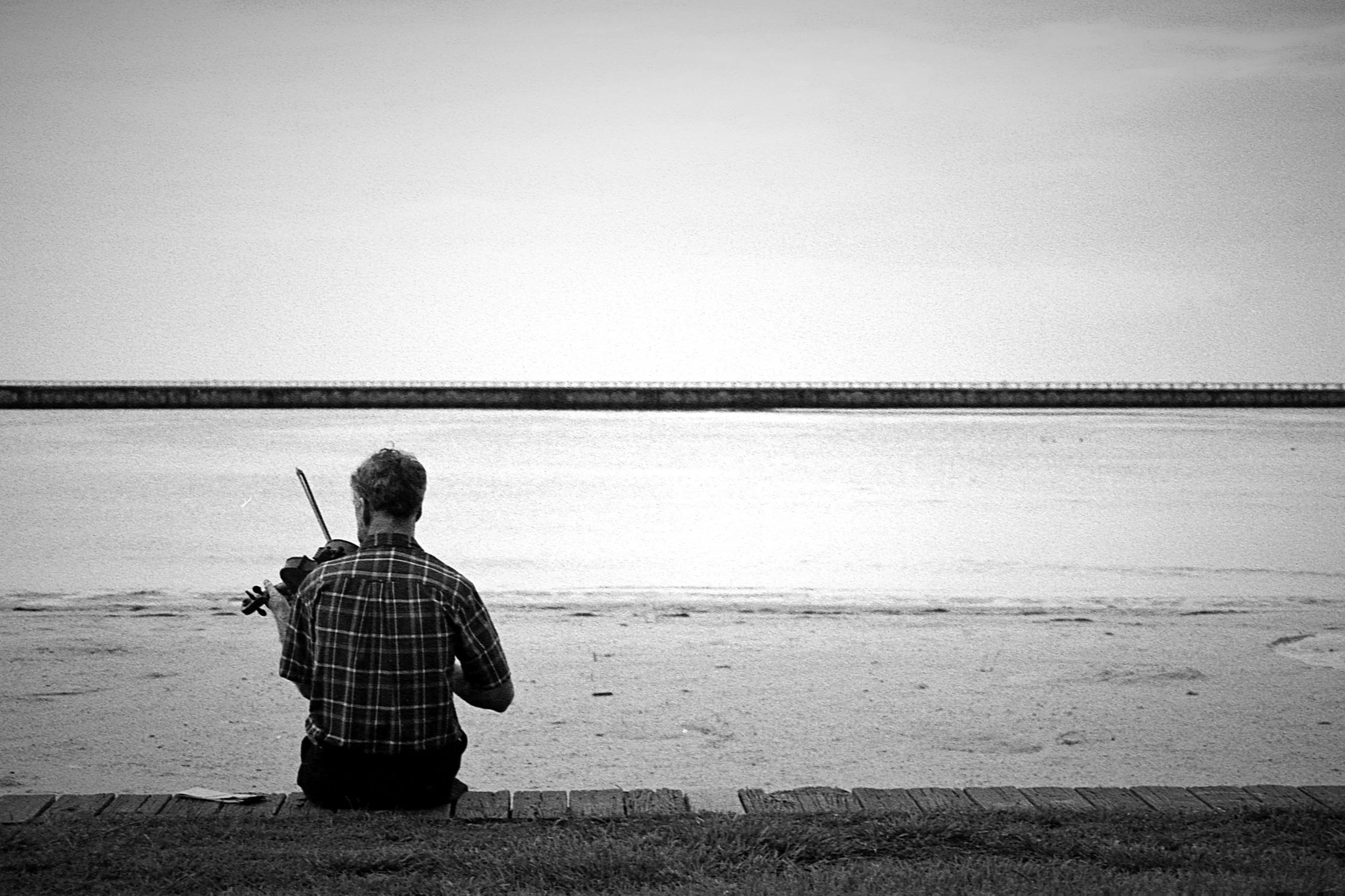A person sitting on a brick border at the beach, playing a violin, with water and a bridge in the background, in black and white.