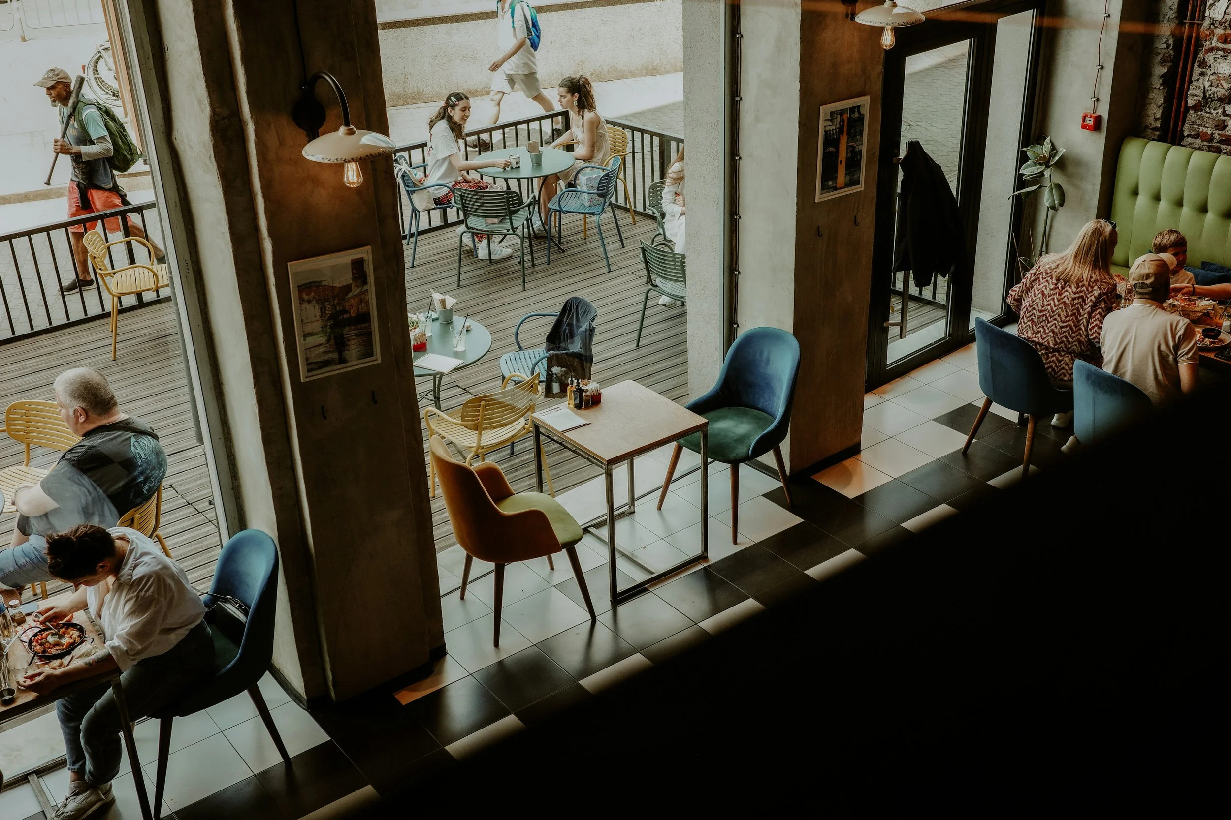 Interior view of a cafe with several groups of people dining and sitting at tables, some outside on a balcony. The cafe features a mix of colorful chairs, tables, exposed brick walls, and large glass windows.
