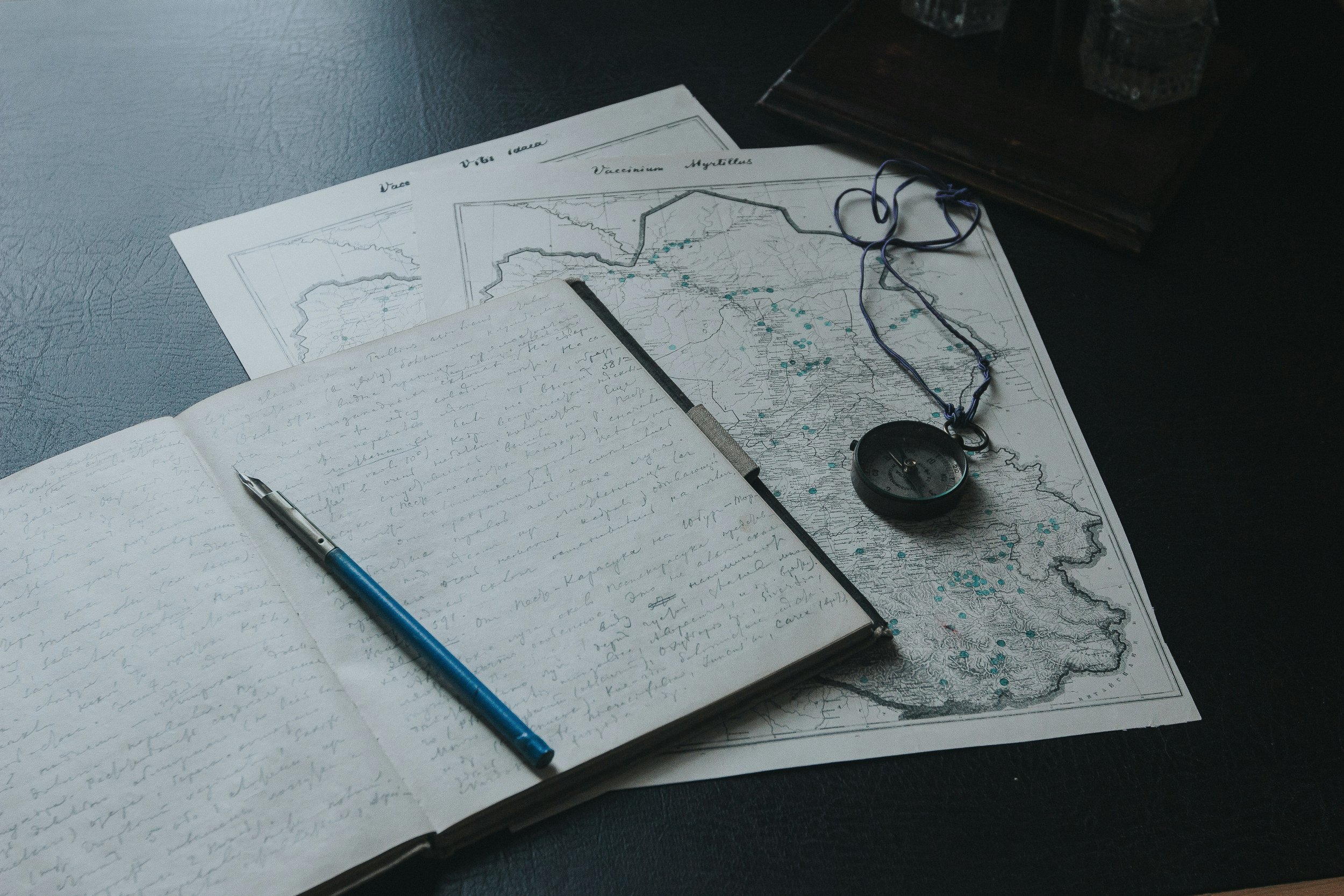 A desk with handwritten notes in an open notebook, a blue pen, a map of Myanmar with marked locations, a compass, and a string. There are additional maps or documents partially visible behind.