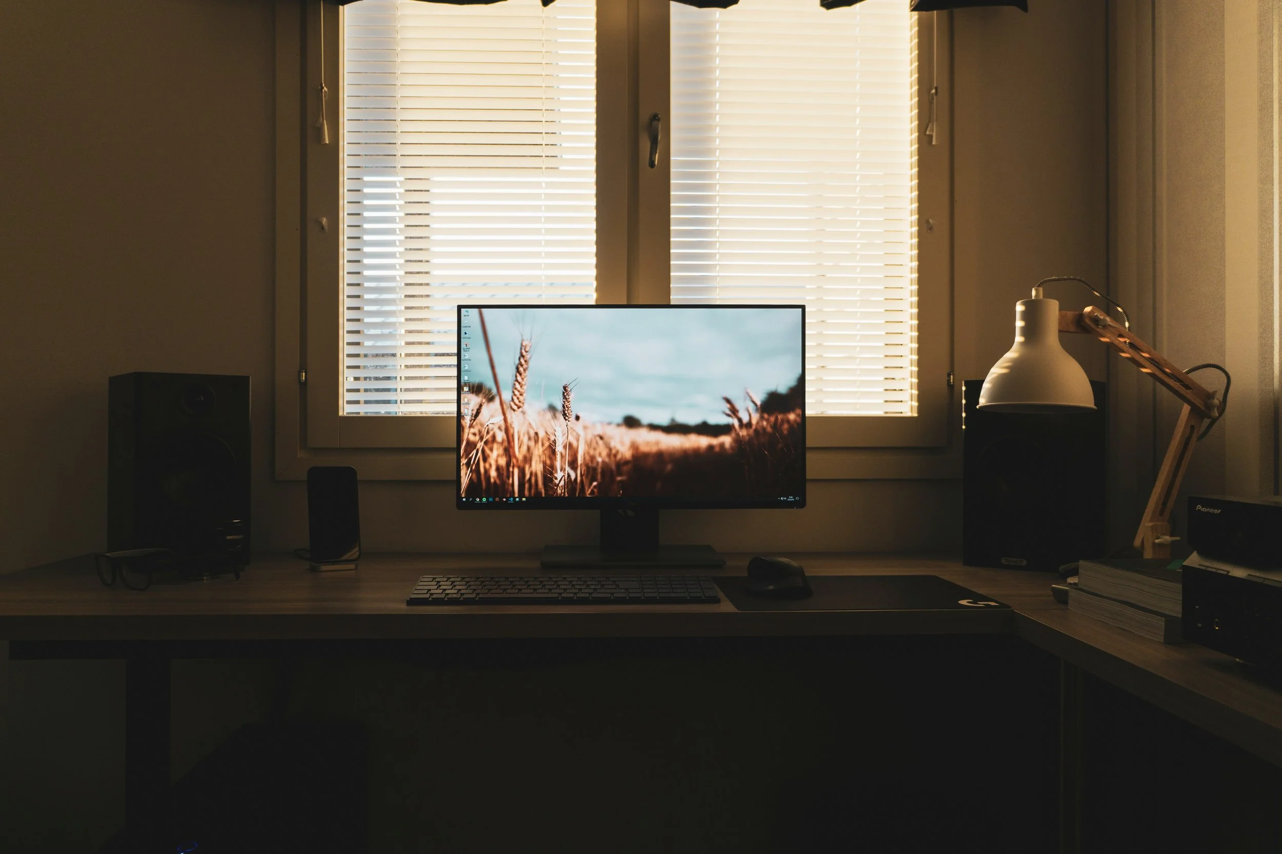 A computer desk setup with a monitor displaying a field of wheat, placed in front of a window with closed blinds, with various desk accessories and devices on the table.
