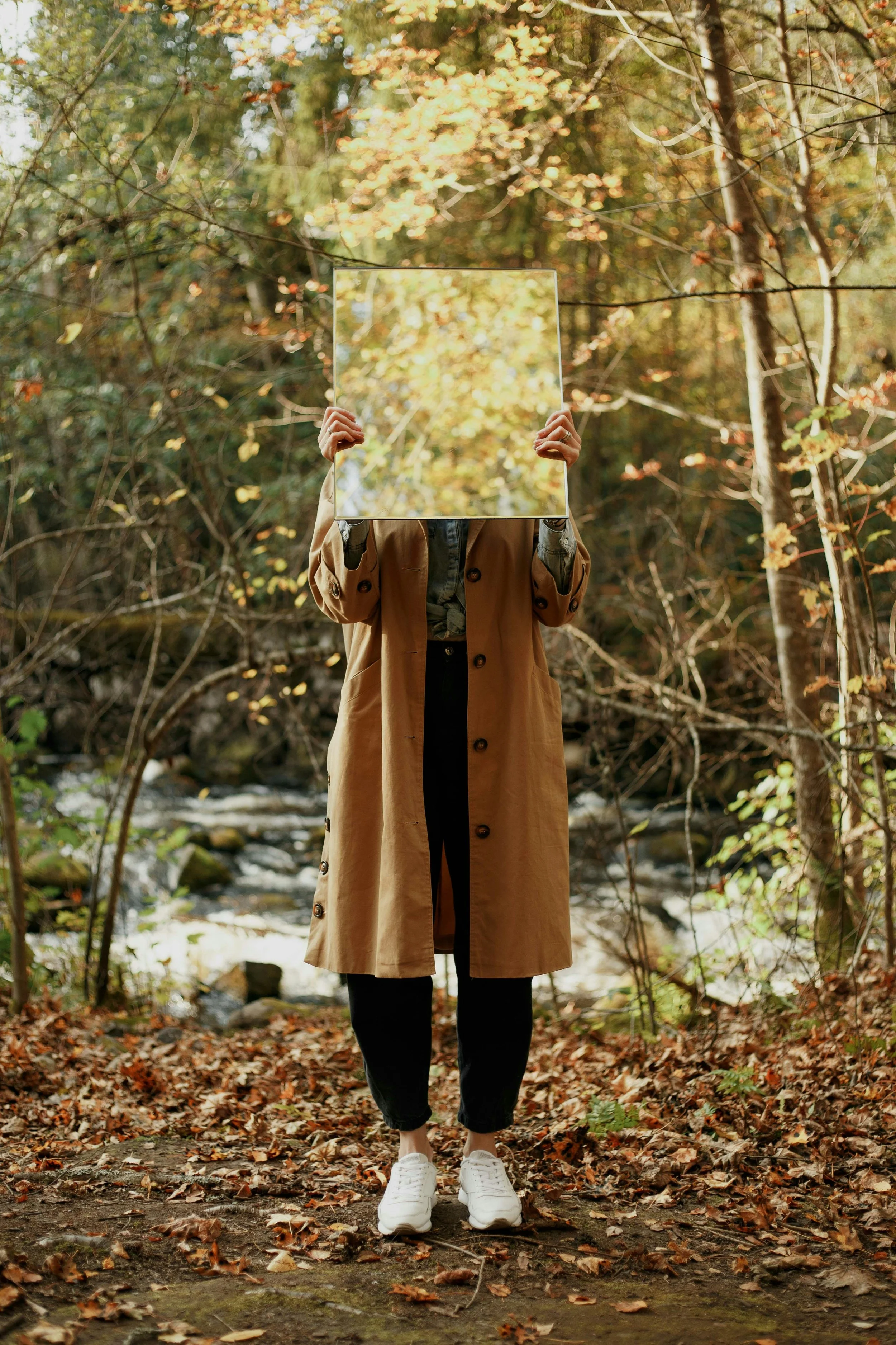 Person standing outdoors in a wooded area, holding a mirror in front of their face that reflects autumn leaves.