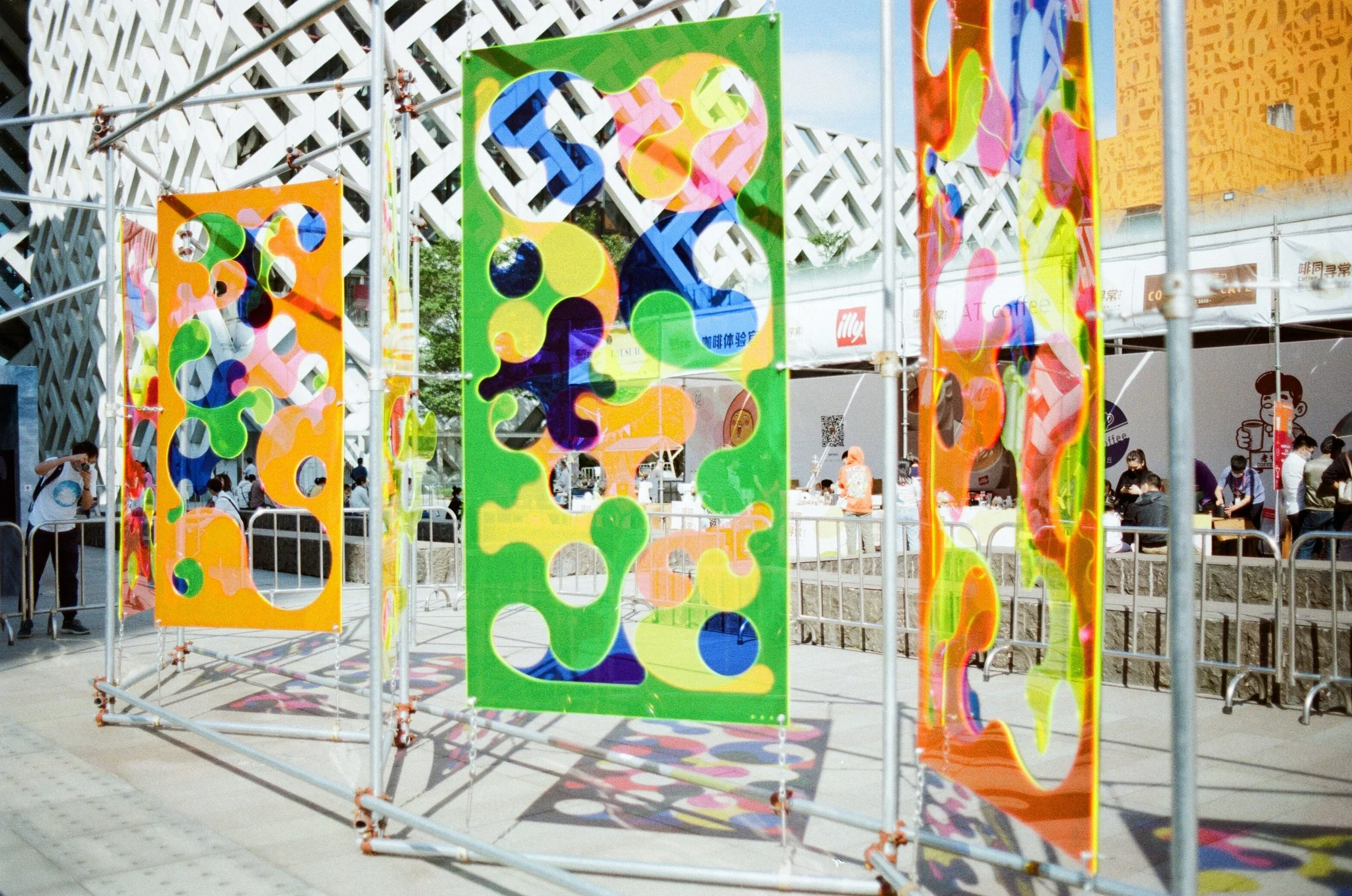 Colorful abstract hanging fabric art installation on a metal frame in an outdoor setting with people walking and a building with lattice architecture in the background.