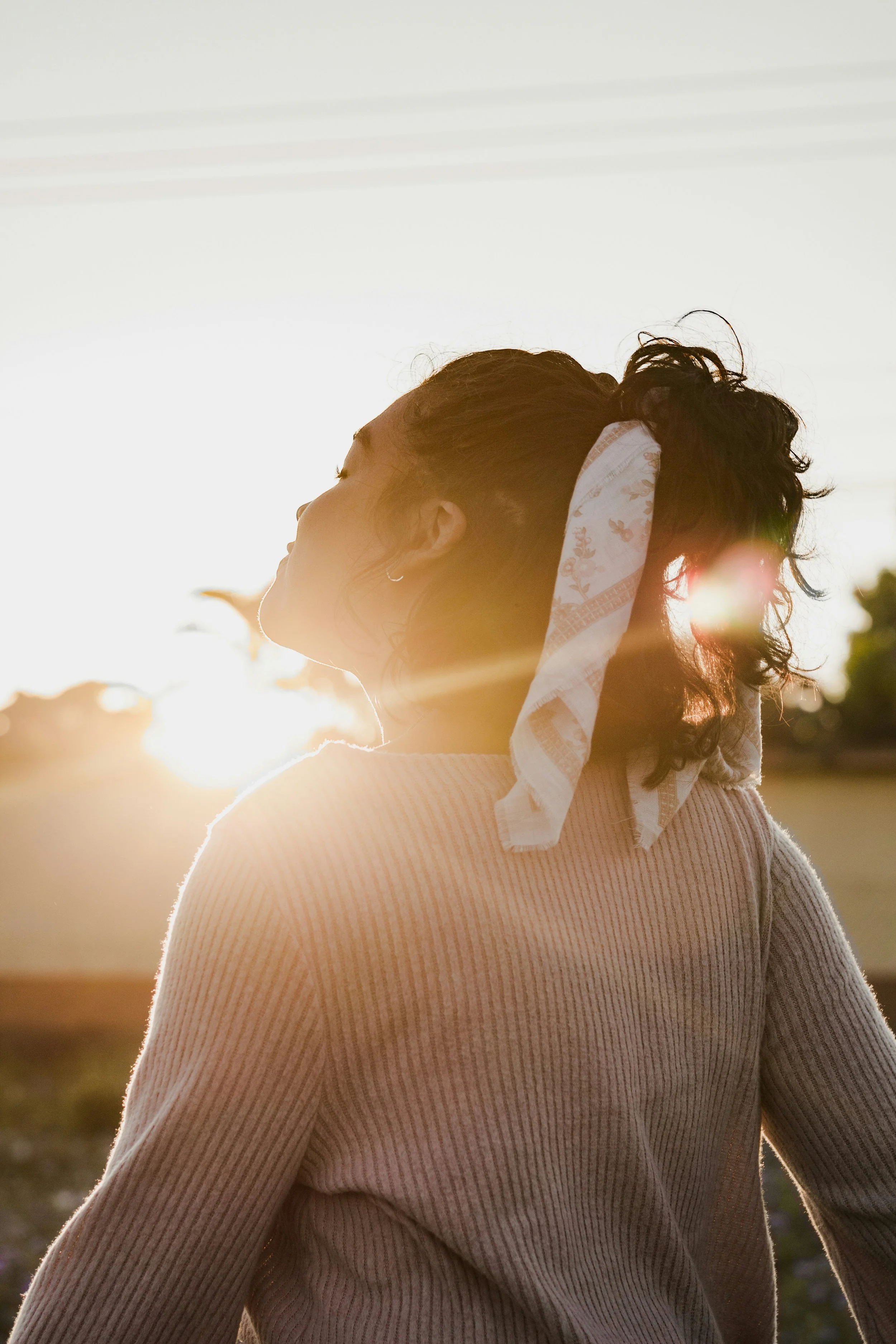 A woman with curly hair tied with a scarf standing outdoors during sunset with the sun shining behind her.