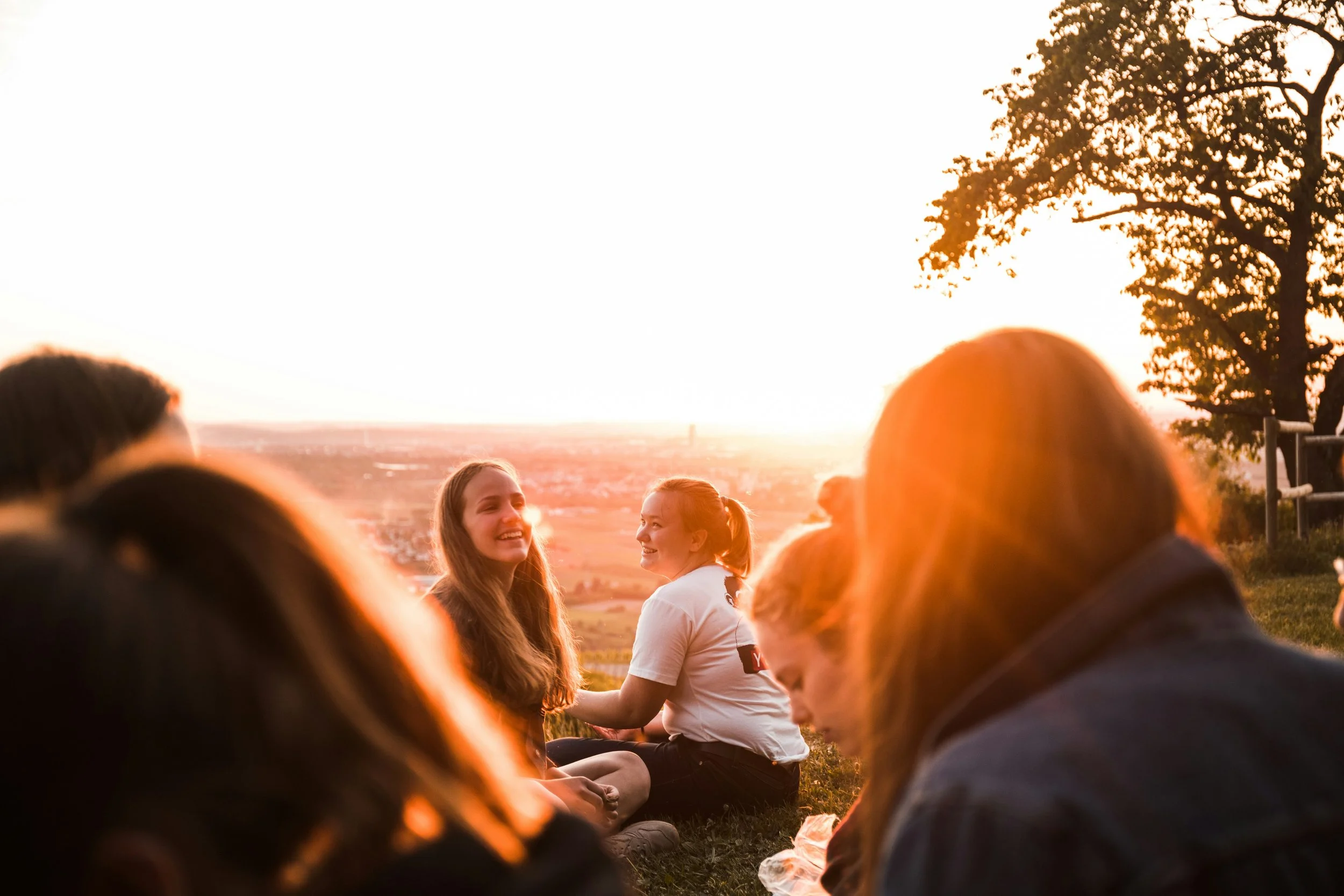 Group of friends sitting outdoors during sunset, smiling and chatting, with a scenic view of the landscape behind them.